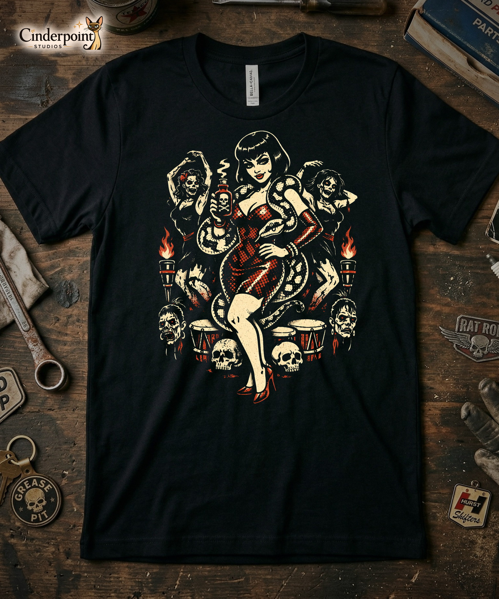 The Voodoo Vixen t-shirt laid flat on a weathered wooden workbench. Surrounded by a vintage 'Hurst Shifters' patch, a 'Rat Rod' wing emblem, an old wrench, and a 'Grease Pit' skull keychain. The high-contrast red and cream art pops against the black fabric and dark wood. Cinderpoint Studios Siamese cat logo is placed in the top-left corner.