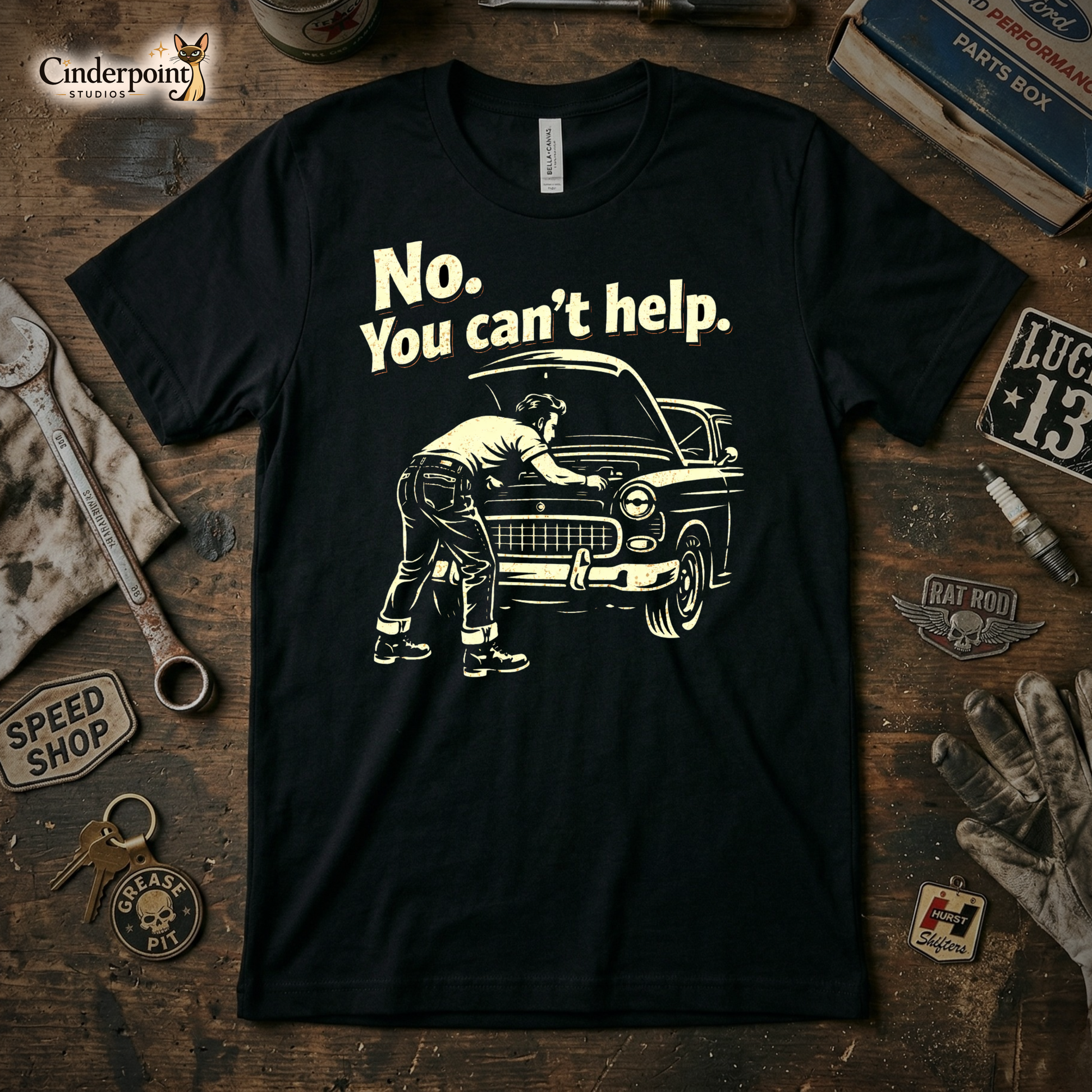 A top-down photograph of a black t-shirt featuring a cream-colored distressed graphic of a man working under a car hood with the text "No. You Can't Help." The shirt is on a rustic wooden workbench surrounded by vintage tools and a speed shop patch.