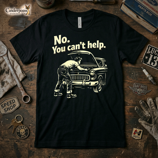 A top-down photograph of a black t-shirt featuring a cream-colored distressed graphic of a man working under a car hood with the text "No. You Can't Help." The shirt is on a rustic wooden workbench surrounded by vintage tools and a speed shop patch.