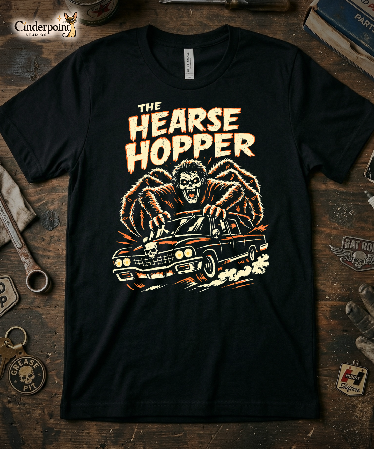 Top-down flat lay of a black Bella Canvas 3001 t-shirt with 'The Hearse Hopper' design on a rustic wooden workshop table with a wrench, spark plug, and vintage car club patches.