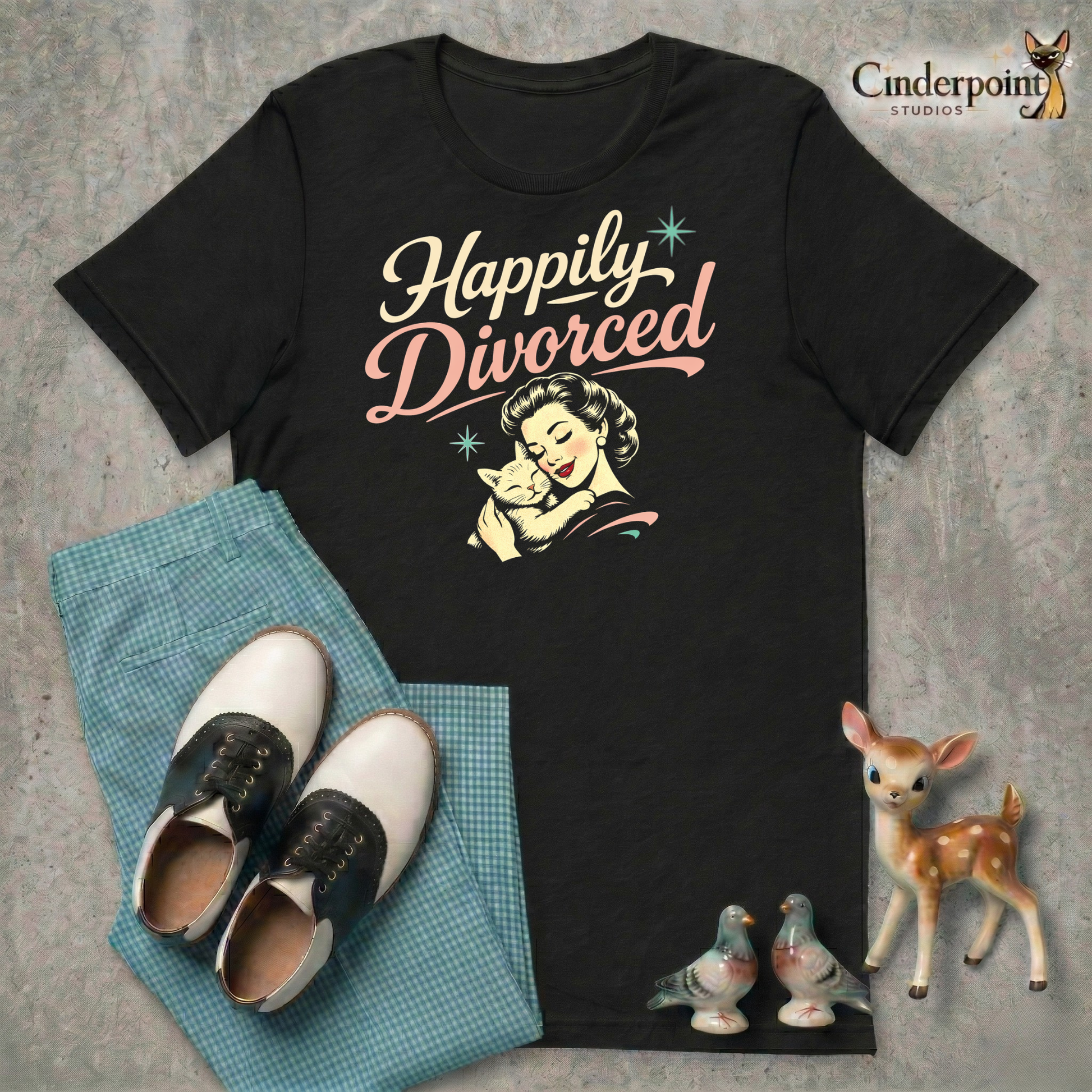 Black t-shirt with 'Happily Divorced' graphic, shoes, and deer figurine on a textured surface.