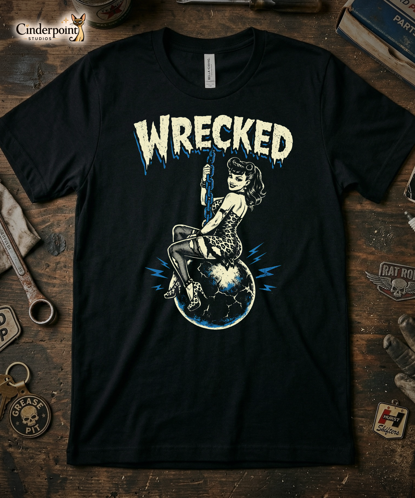 A premium black t-shirt featuring the 'Wrecked' design. A stippled, retro-style pinup girl in a leopard-print dress and fishnets sits atop a cracked, electric-blue globe suspended by a heavy blue chain. Distressed 'WRECKED' typography arcs over the top in bone-cream. The vertical print is centered high on the chest for a boutique look.