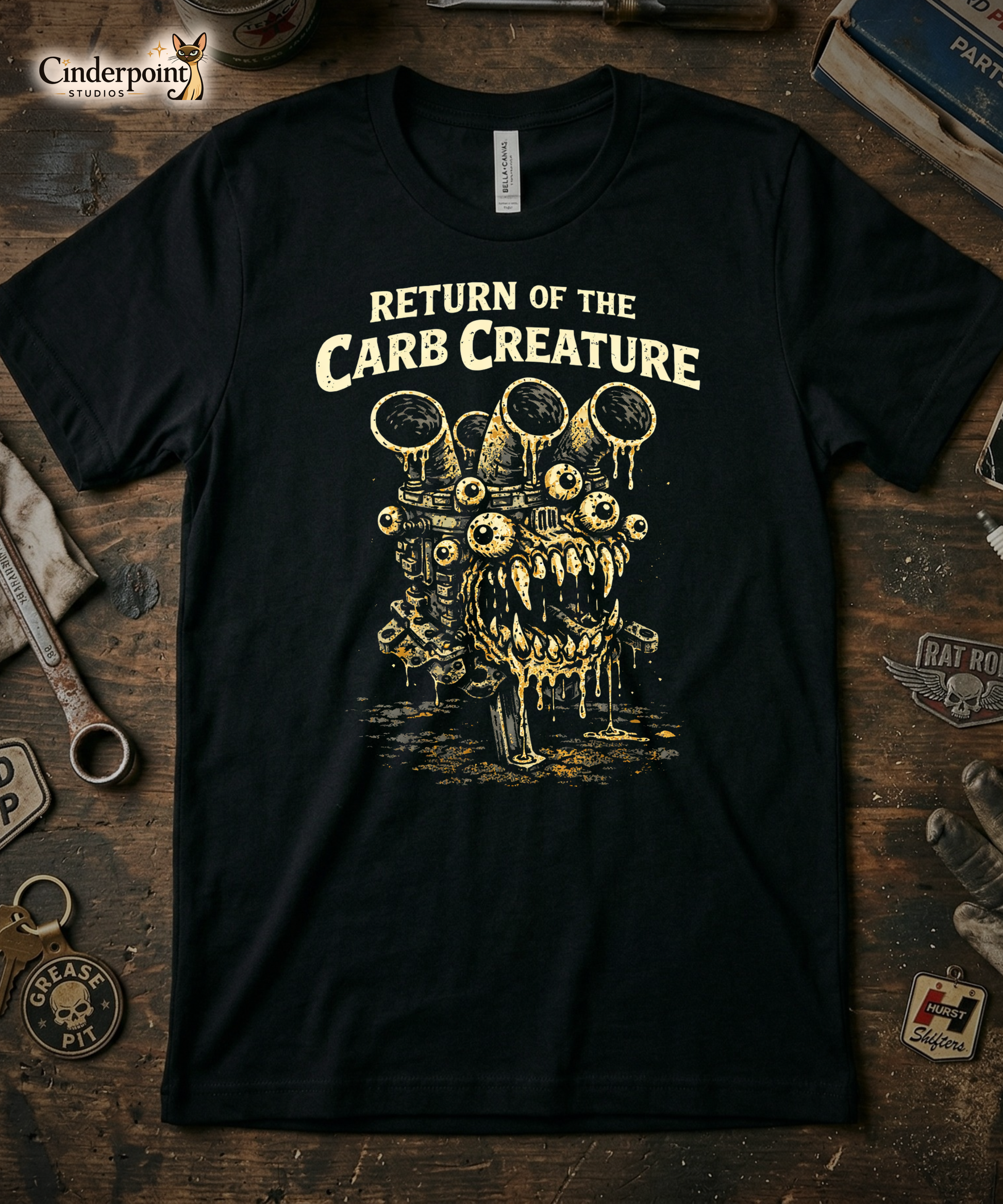 Top-down flat lay of a black Bella Canvas 3001 t-shirt with the 'Return of the Carb Creature' many-eyed engine monster design on a rustic wooden workshop table with a wrench, spark plug, and vintage car club patches.