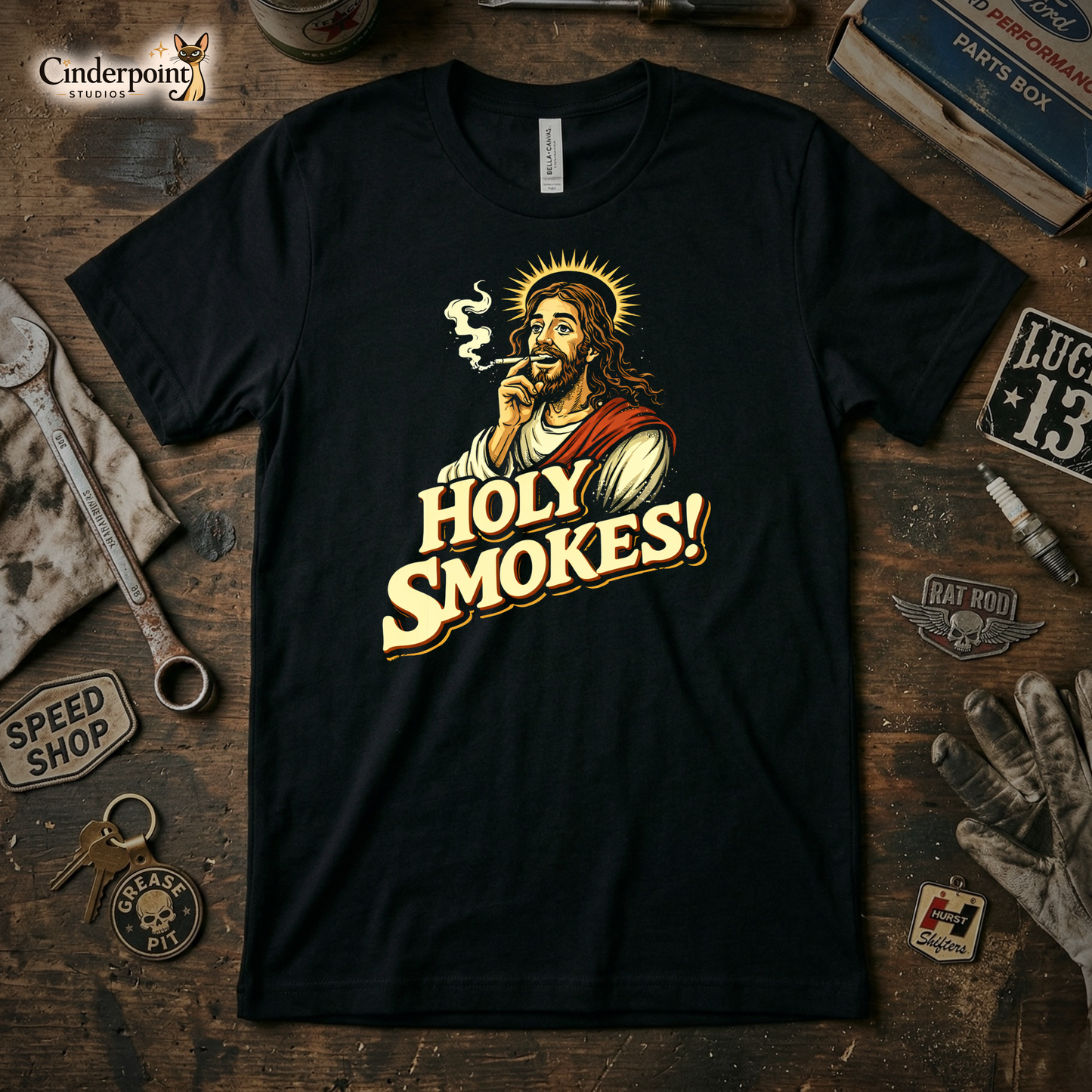 A top-down photograph of a black t-shirt with a distressed graphic of a haloed Jesus smoking a cigarette above the text "HOLY SMOKES!" The shirt is centered on a rustic wooden workbench with vintage tools and a greasy rag nearby.