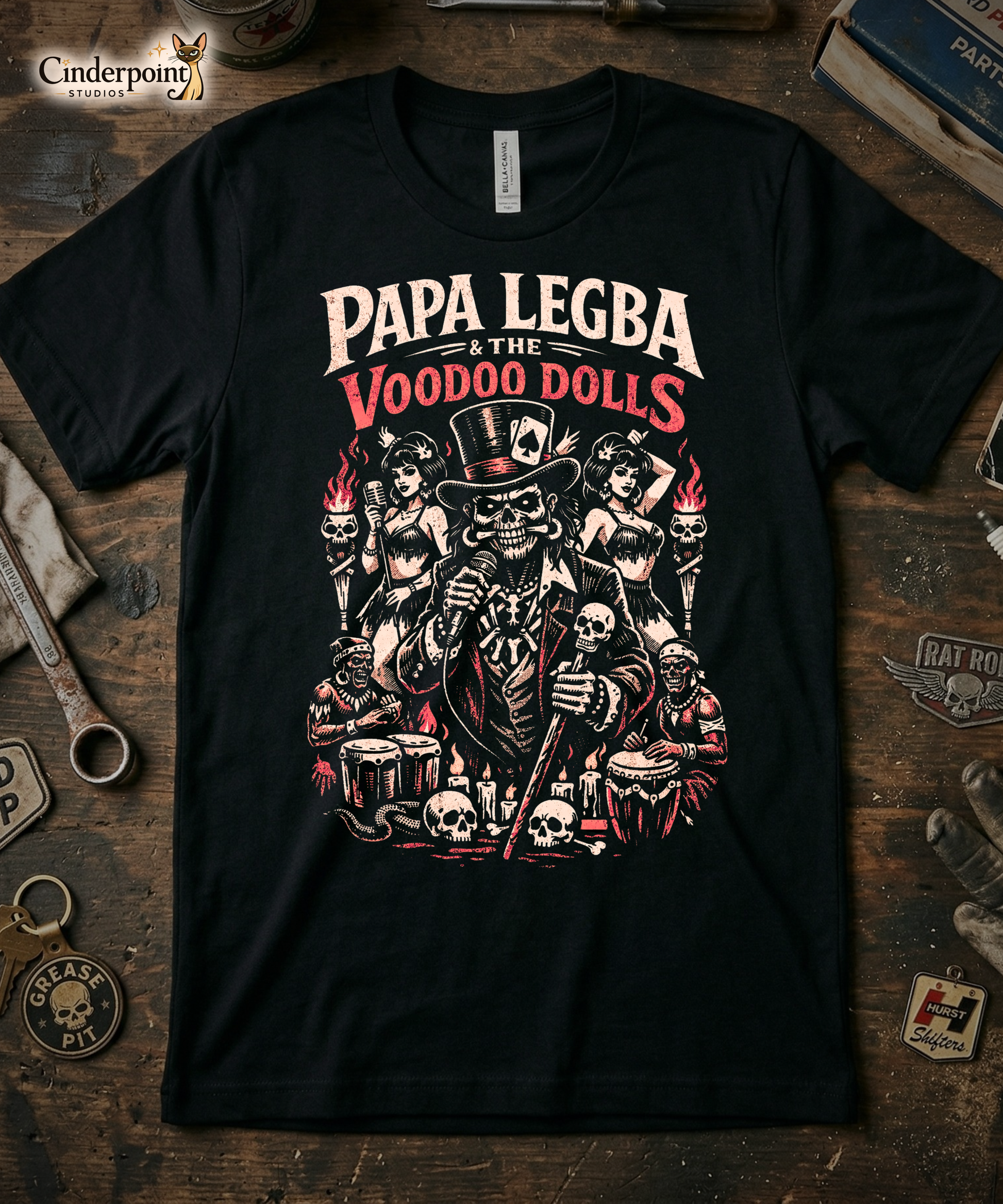 A rugged product mockup of the Papa Legba t-shirt laid flat on a distressed wooden workshop bench. The detailed retro psychobilly art is surrounded by period-correct tools, an 'Hurst Shifters' vintage patch, a 'Rat Rod' wing emblem, and a 'Grease Pit' skull keychain. The top left corner features the Cinderpoint Studios Siamese cat logo.