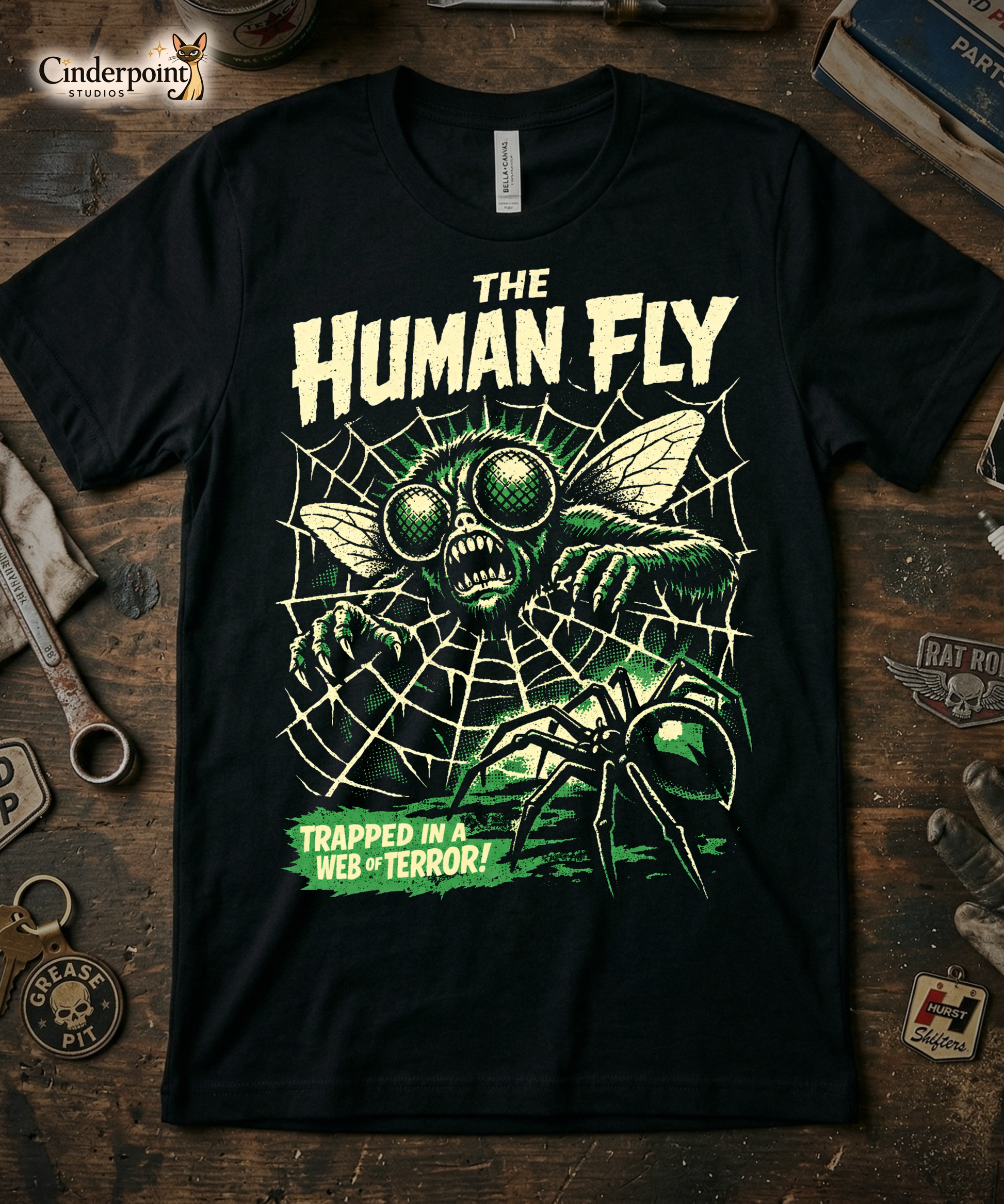 'The Human Fly' t-shirt laid flat on a weathered wooden workbench. Surrounding the shirt are a 'Grease Pit' skull keychain, a 'Hurst Shifters' patch, and vintage automotive parts.