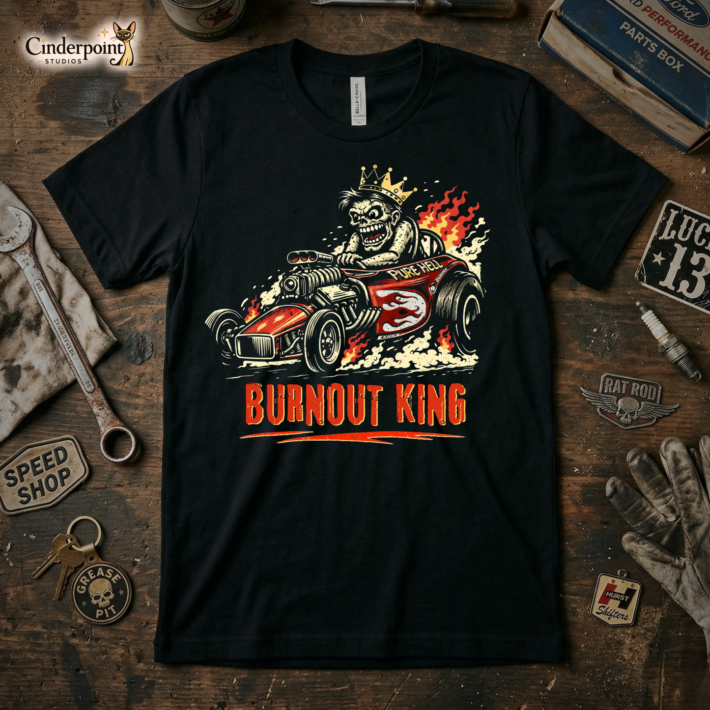 A black t-shirt featuring a vibrant, distressed "Burnout King" graphic with a skeleton driving a flame-throwing dragster. The shirt is laid on a wooden workbench with a vintage wrench, spark plugs, and a "Speed Shop" patch.