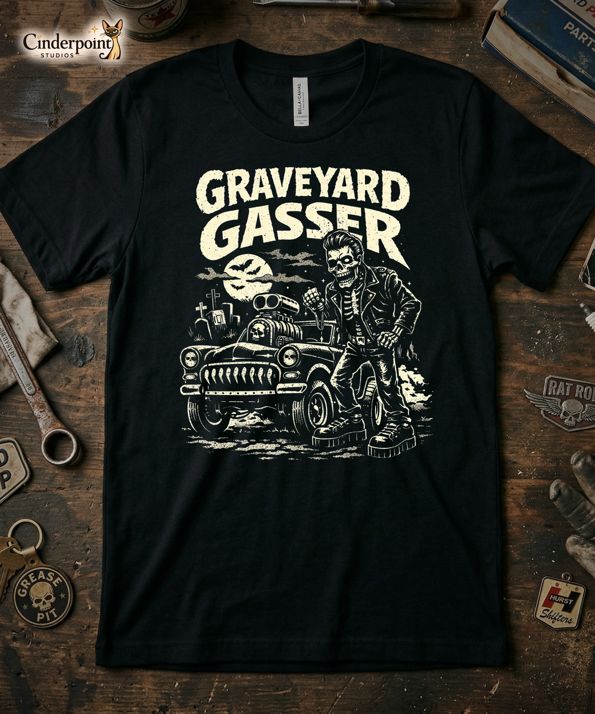 Top-down flat lay of a black Bella Canvas 3001 t-shirt with the 'Graveyard Gasser' skeleton mechanic design on a rustic wooden workshop table with a wrench, spark plug, and vintage car club patches.