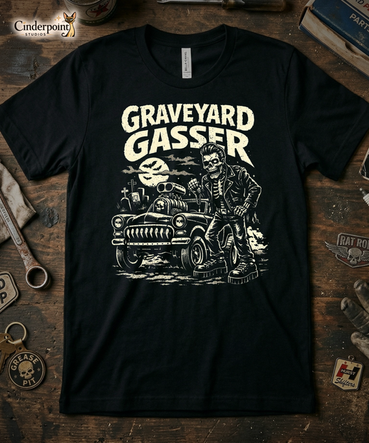 Top-down flat lay of a black Bella Canvas 3001 t-shirt with the 'Graveyard Gasser' skeleton mechanic design on a rustic wooden workshop table with a wrench, spark plug, and vintage car club patches.
