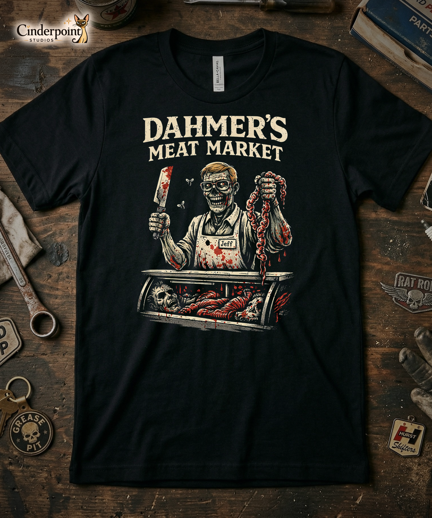 Top-down flat lay of a black Bella Canvas 3001 t-shirt with the 'Dahmer's Meat Market' zombie butcher design on a rustic wooden workshop table with a wrench, spark plug, and vintage car club patches.
