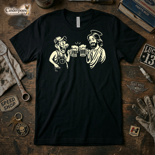 A top-down photograph of a black t-shirt featuring a cream-colored cartoon graphic of a man in overalls clinking beer mugs with Jesus Christ. The shirt is centered on a rustic wooden workbench surrounded by vintage tools and a "Speed Shop" patch.