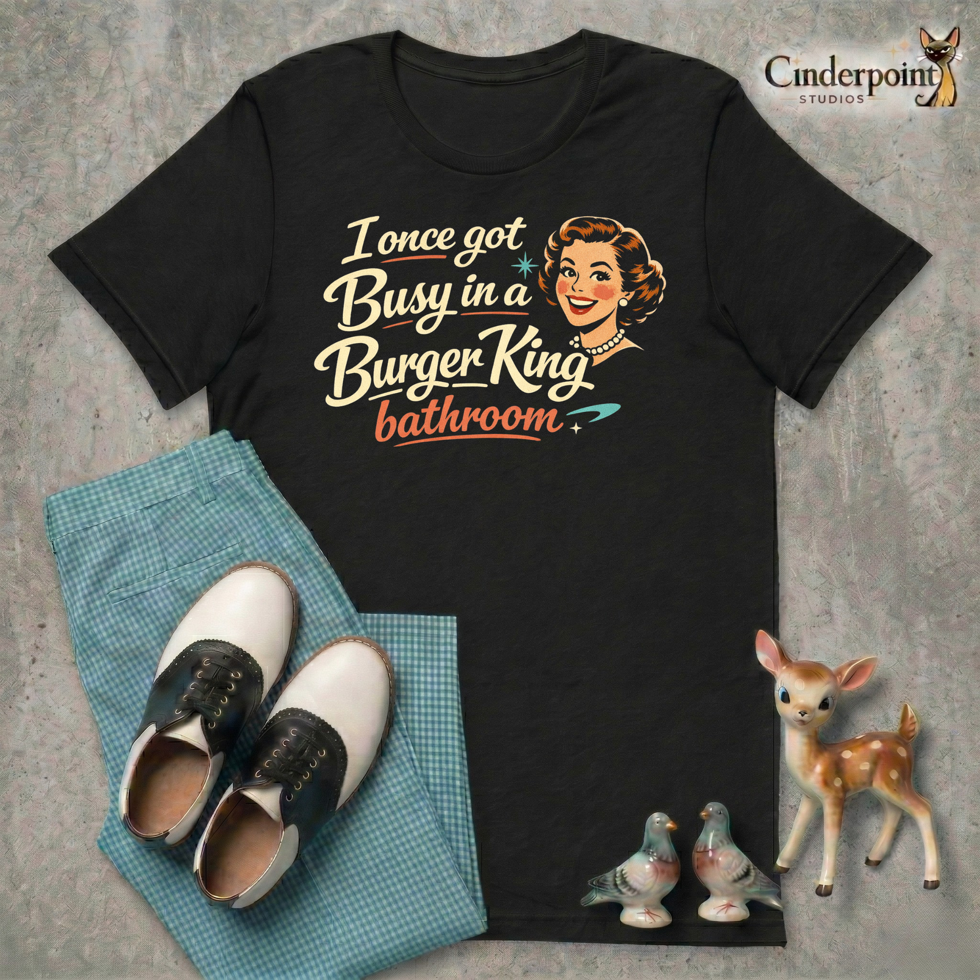 Flat layout of a black t-shirt with a 1950s housewife illustration that reads, "I once got busy in a Burger King Bathroom"