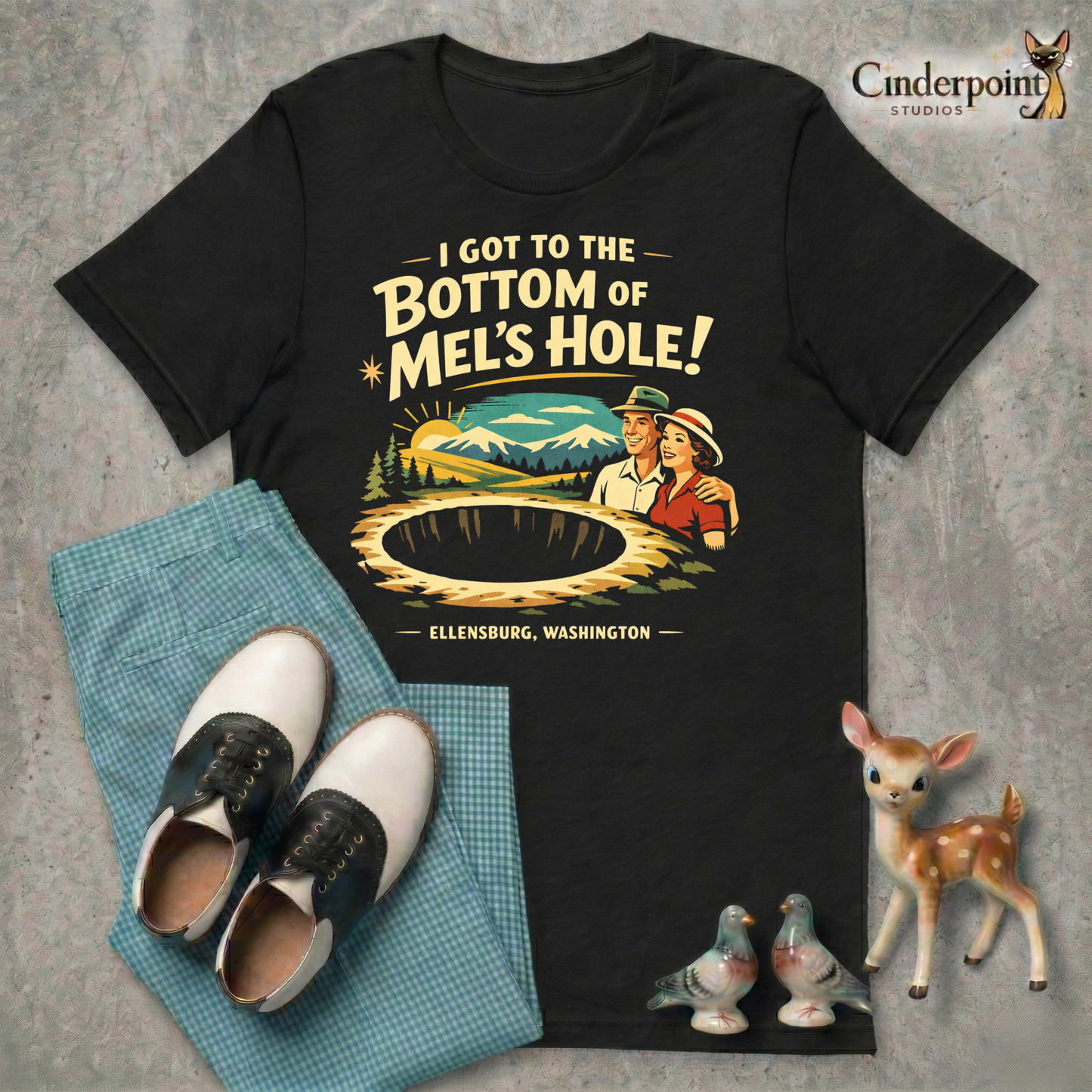 Black retro graphic t-shirt featuring a vintage travel-style illustration of smiling tourists beside a giant mysterious hole with the phrase I Got to the Bottom of Mel’s Hole and Ellensburg, Washington.