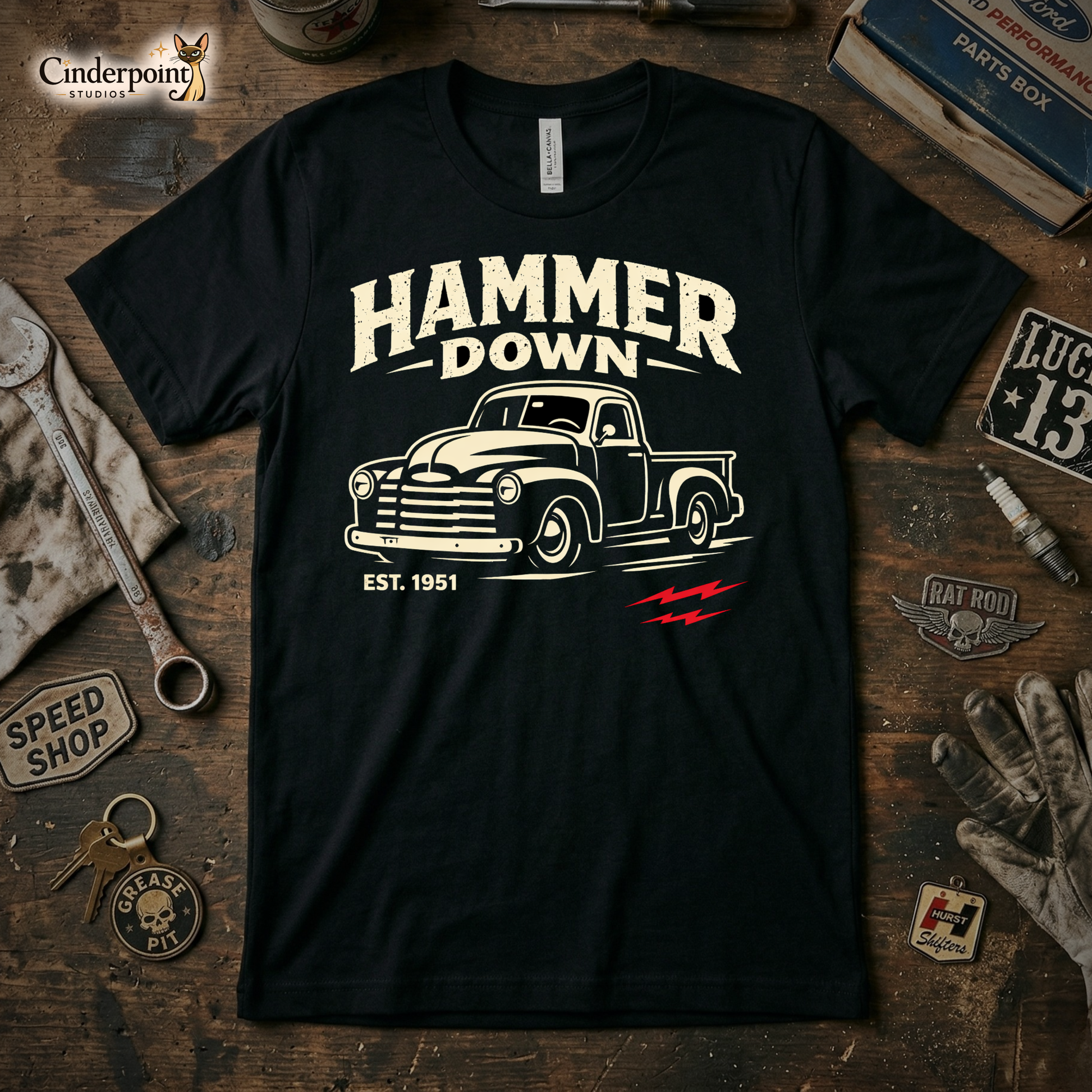 A top-down photograph of a black t-shirt featuring a distressed "Hammer Down" 1951 Chevy truck graphic. The shirt is on a rustic wooden workbench surrounded by vintage tools, a greasy rag, and a "Speed Shop" patch.