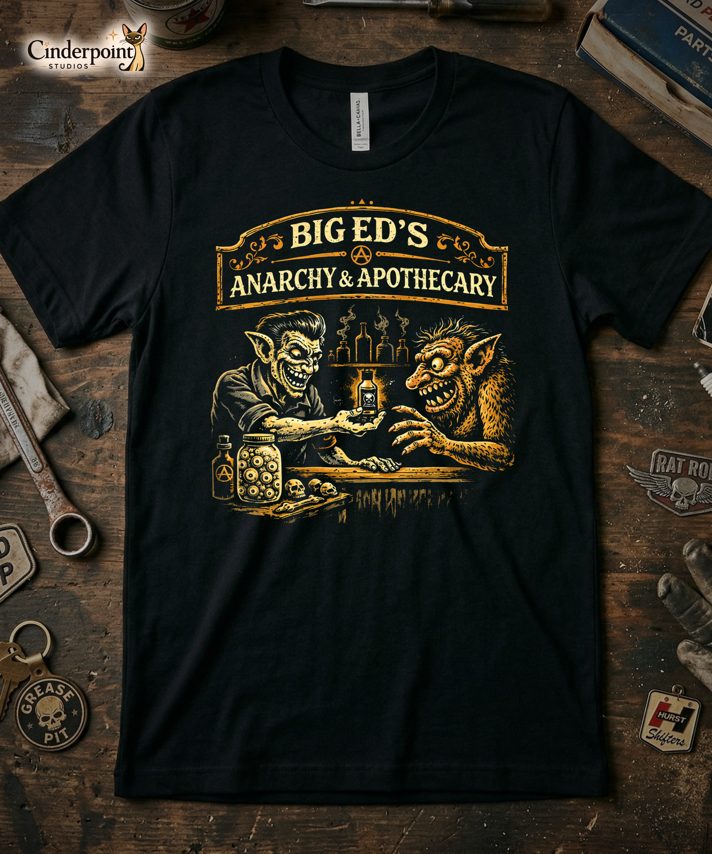 Top-down flat lay of a black Bella Canvas 3001 t-shirt with 'Big Ed's Anarchy & Apothecary' design on a rustic wooden workshop table with a wrench, spark plug, and vintage car club patches.