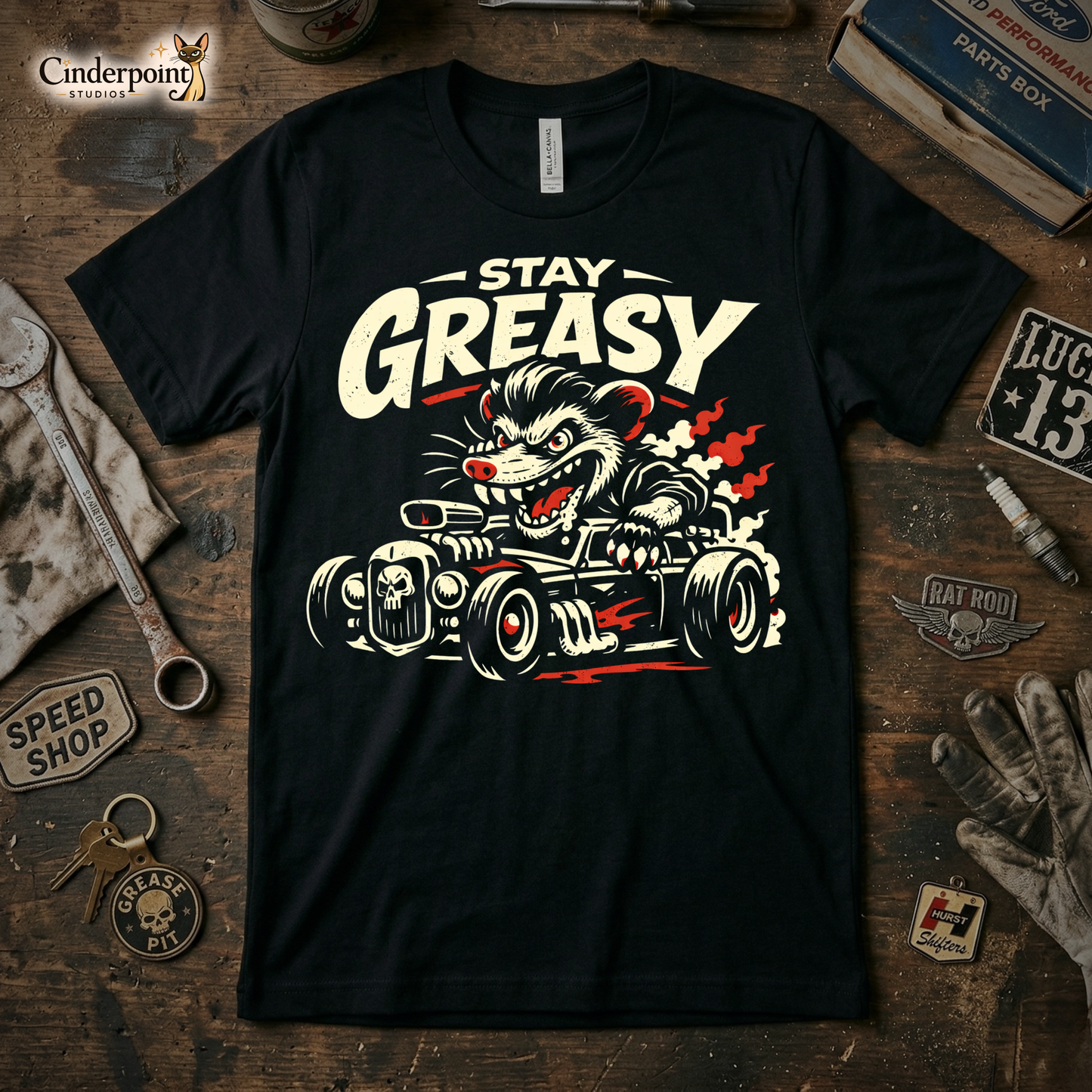 A black t-shirt featuring a distressed "Stay Greasy" rat rod graphic, surrounded by vintage tools, a greasy rag, and car parts on a wooden workbench.