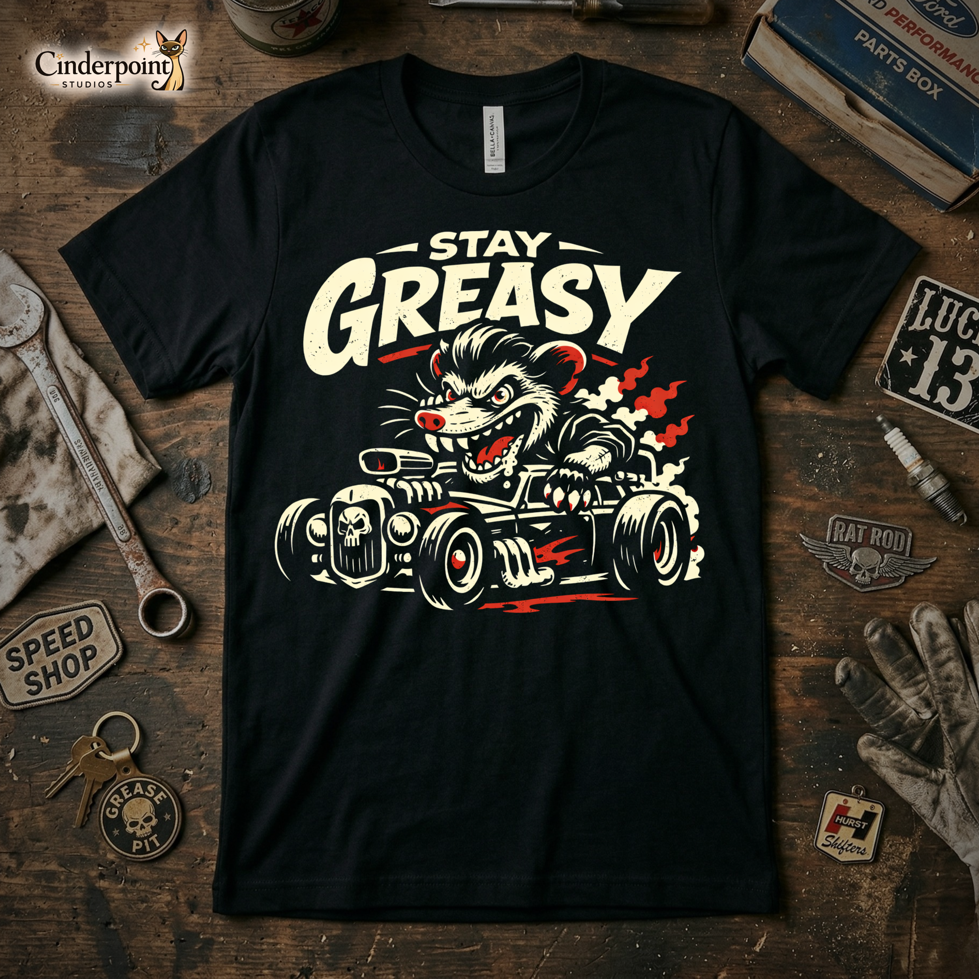 A black t-shirt featuring a distressed "Stay Greasy" rat rod graphic, surrounded by vintage tools, a greasy rag, and car parts on a wooden workbench.