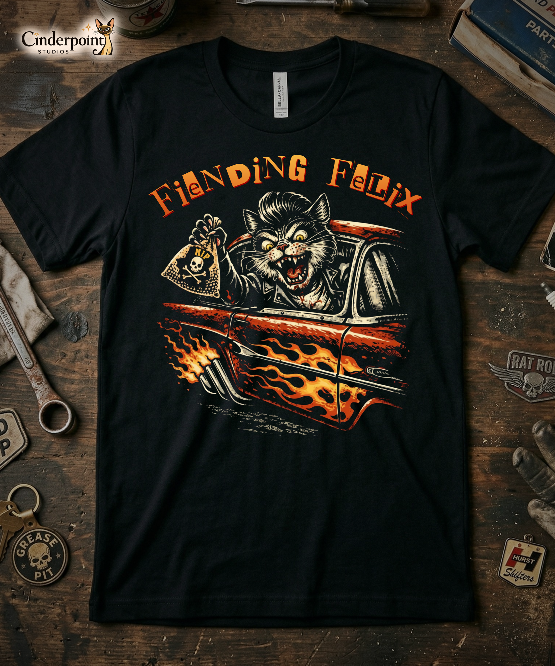 Top-down flat lay of a black Bella Canvas 3001 t-shirt with the 'Fiending Felix' hot rod cat design on a rustic wooden workshop table with a wrench, spark plug, and vintage car club patches.