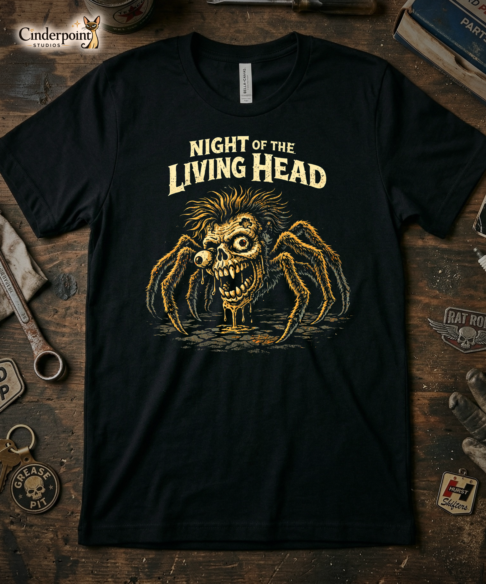 Top-down flat lay of a black Bella Canvas 3001 t-shirt with 'Night of the Living Head' spider zombie design on a rustic wooden workshop table with a wrench, spark plug, and vintage car club patches.