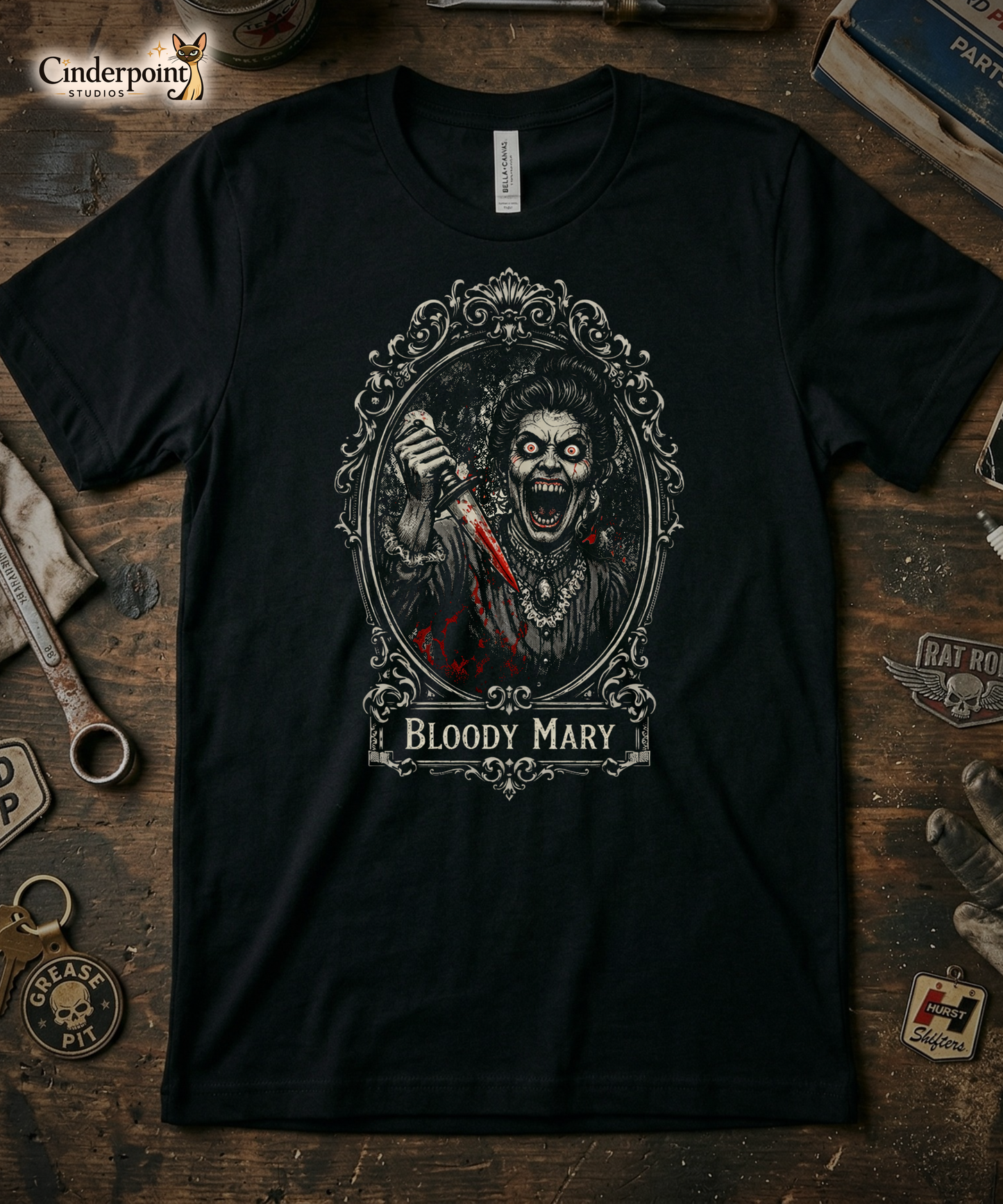 Top-down flat lay of a black Bella Canvas 3001 t-shirt with the 'Bloody Mary' Victorian Goth monster mirror design on a rustic wooden workshop table with a wrench, spark plug, and vintage car club patches.