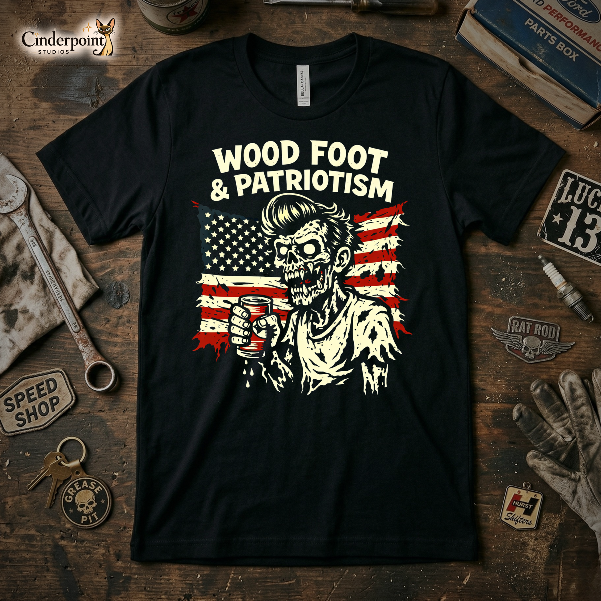 A black t-shirt featuring a distressed cream and red graphic of a zombie holding a beverage in front of a tattered American flag with the text "WOOD FOOT & PATRIOTISM." The shirt is on a rustic wooden workbench surrounded by vintage tools and speed shop badges.