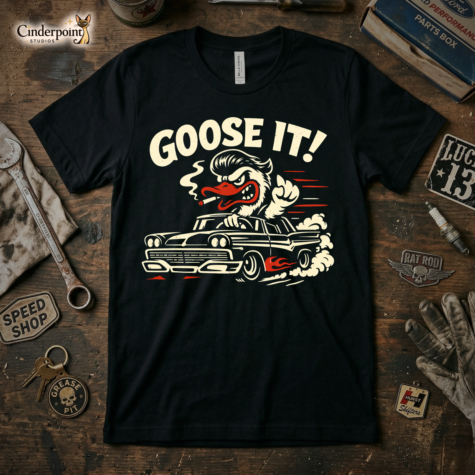 A top-down photograph of a black t-shirt featuring a distressed "Goose It!" graphic of a greaser goose driving a flaming hot rod. The shirt is on a rustic wooden workbench surrounded by vintage tools, keys, and a speed shop patch.