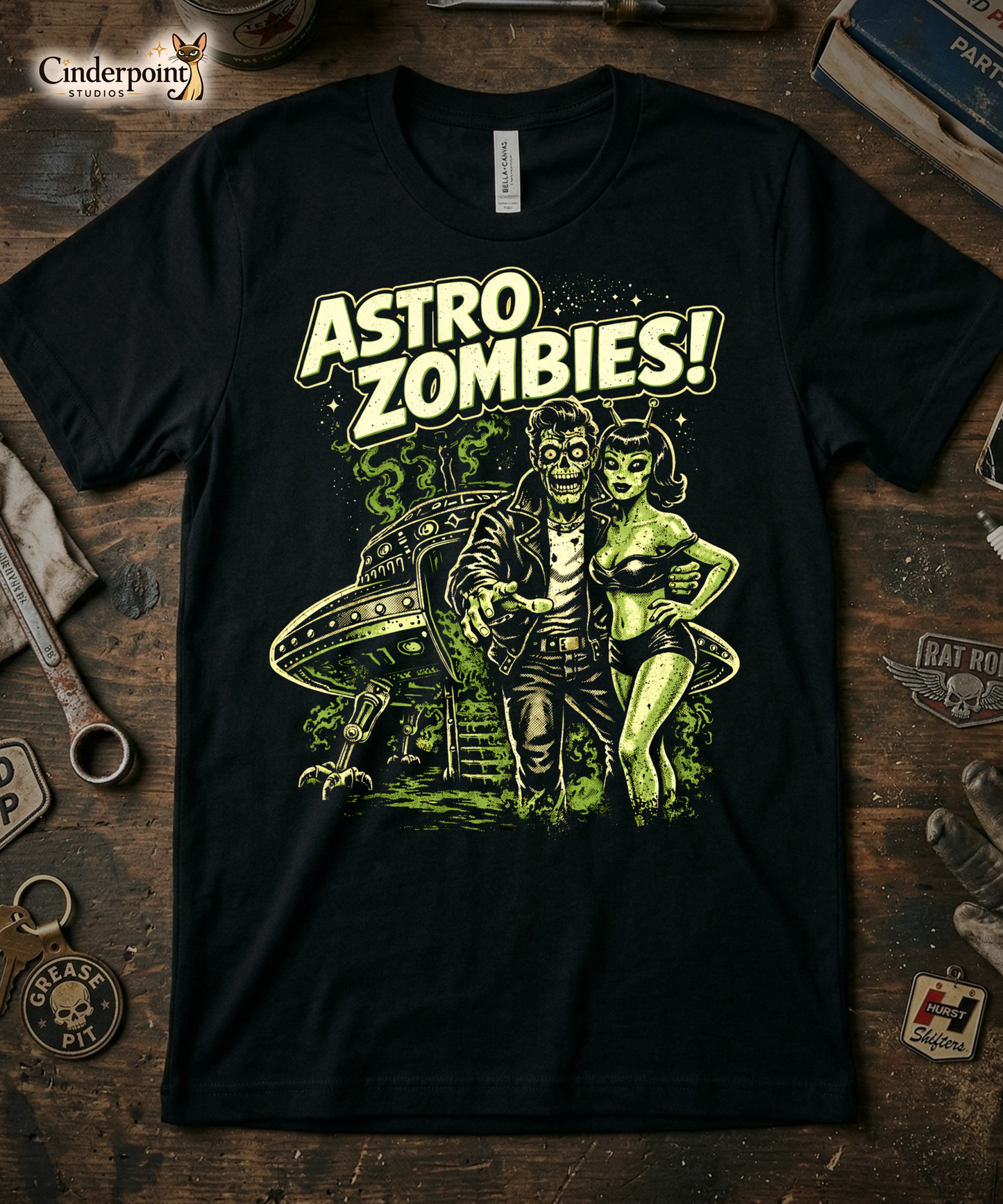 'Astro Zombies!' t-shirt laid flat on a weathered wooden workbench, styled with a 'Grease Pit' keychain, vintage tools, and a 'Hurst Shifters' patch.