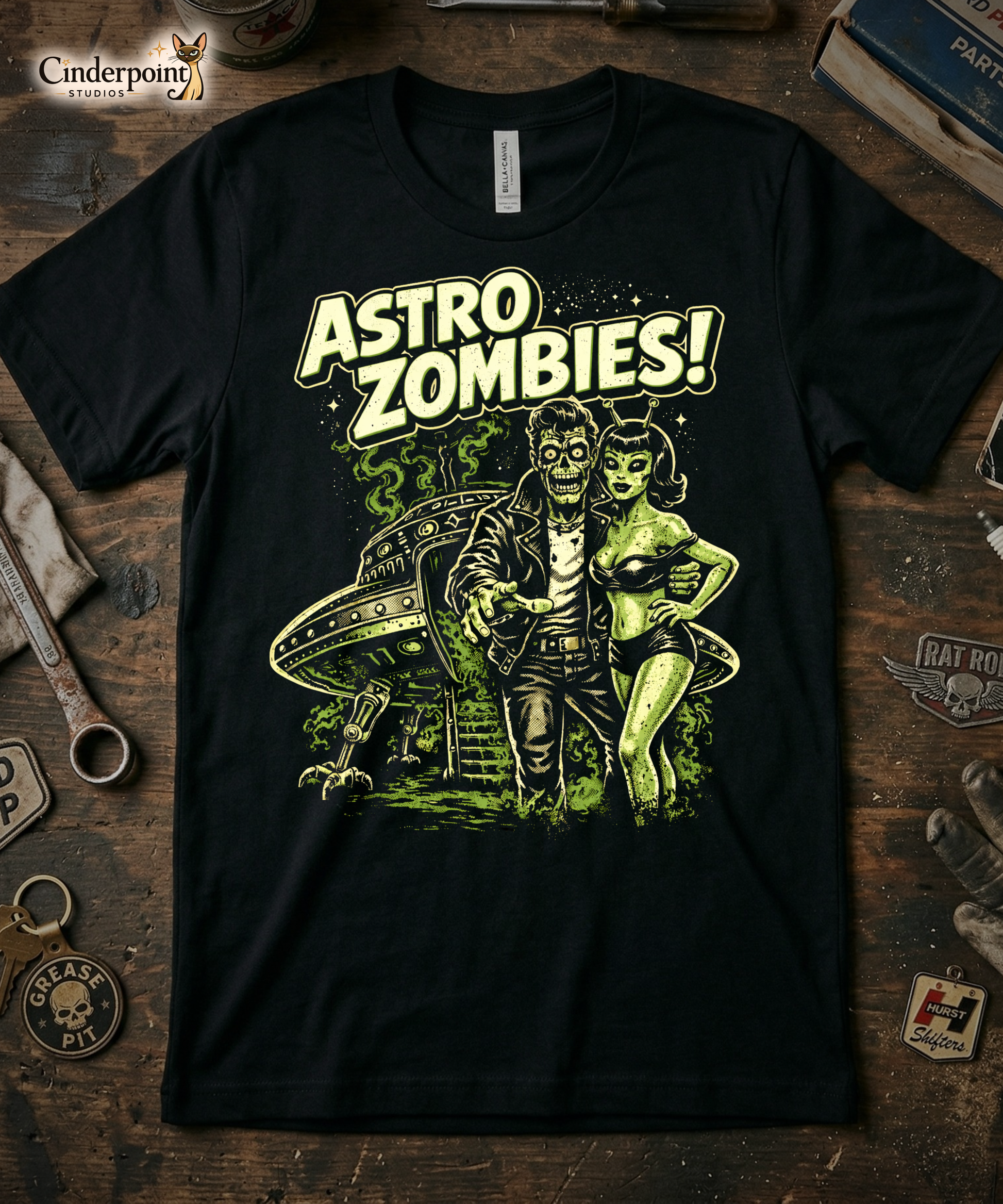 'Astro Zombies!' t-shirt laid flat on a weathered wooden workbench, styled with a 'Grease Pit' keychain, vintage tools, and a 'Hurst Shifters' patch.