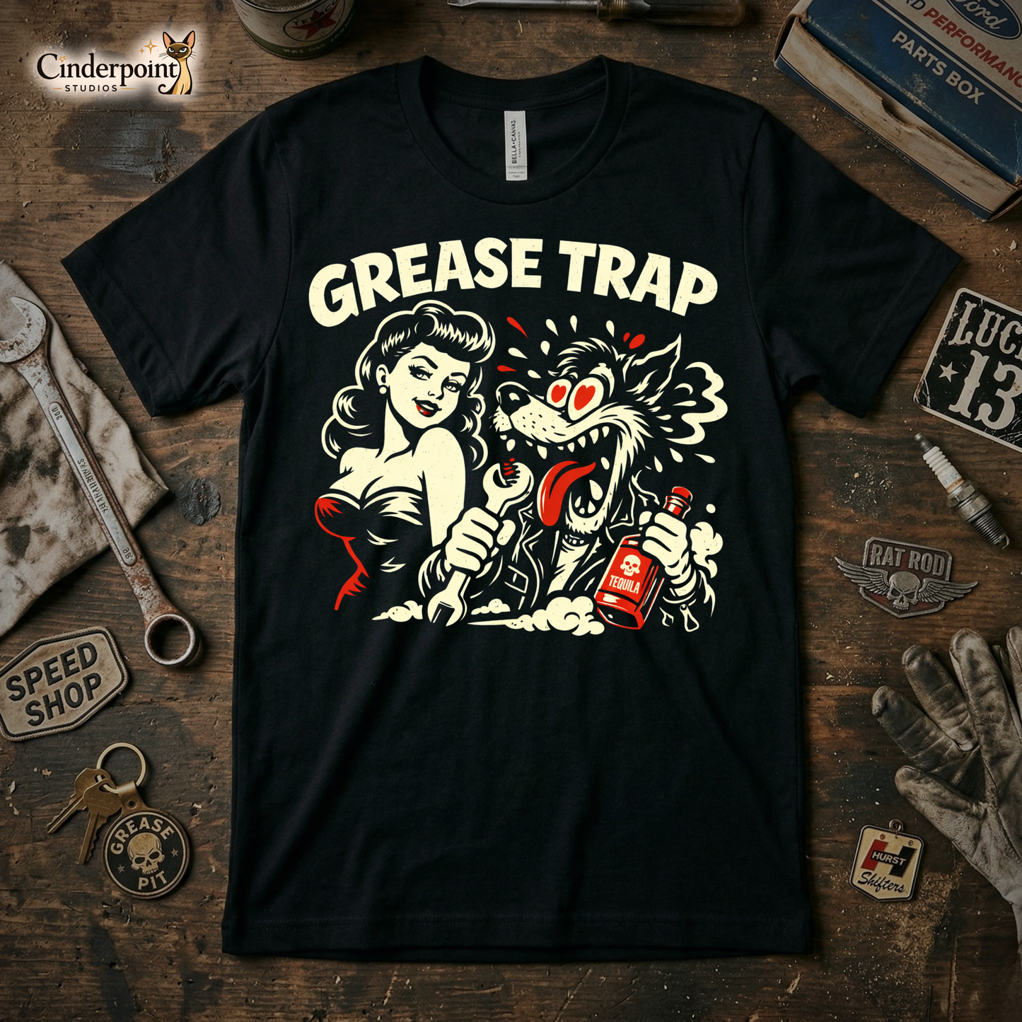 A black t-shirt featuring a distressed "Grease Trap" graphic with a classic pin-up and a manic wolf, surrounded by vintage tools and car parts on a weathered wooden workbench.