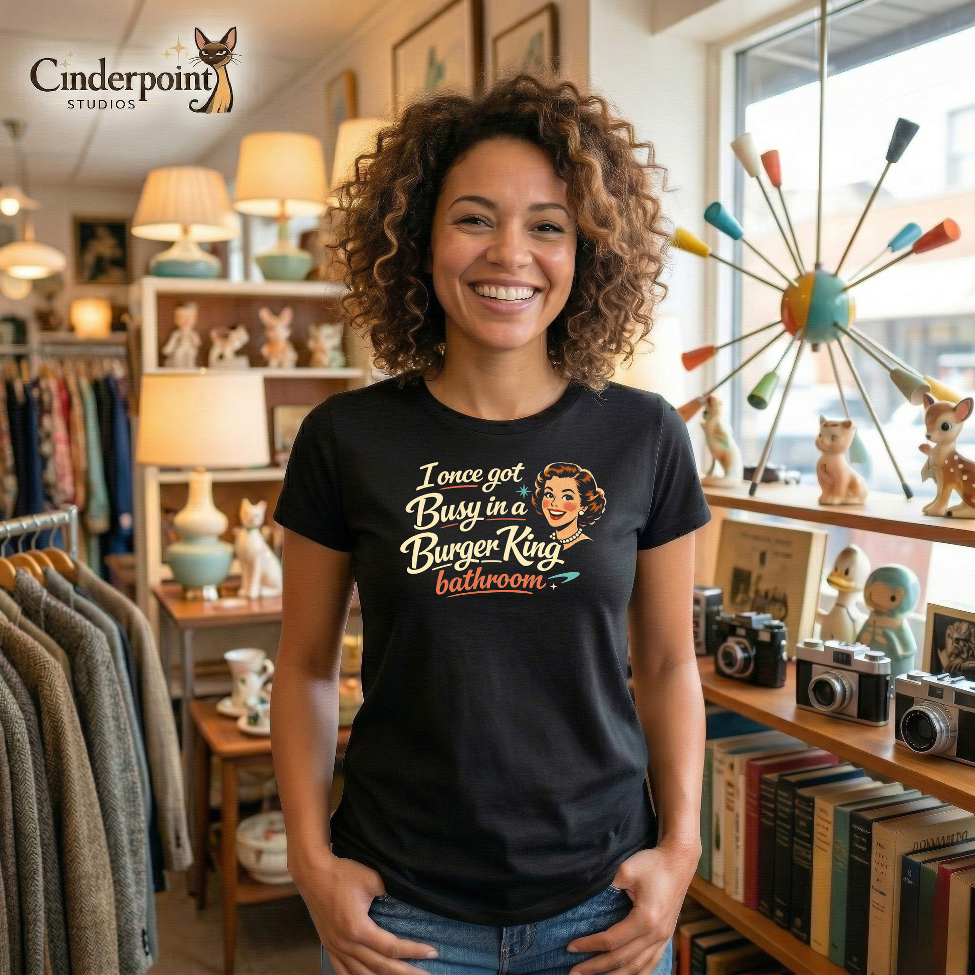 A lifestyle shot of a pretty woman with curly hair in a vintage store. She is smiling proudly and wearing a black t-shirt with a smiling 1950s housewife illustration that reads: "I once got busy in a burger king bathroom."