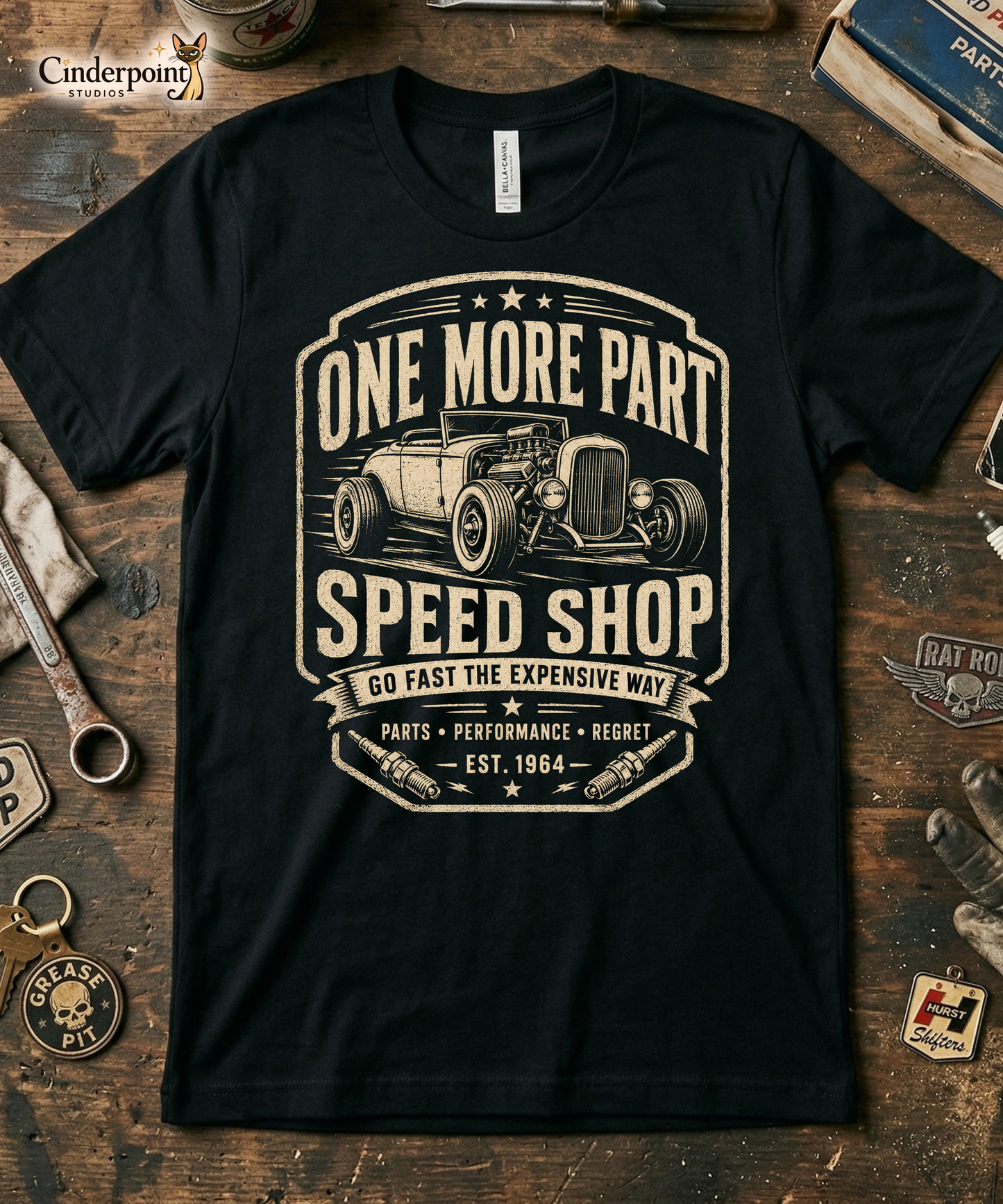 Workbench with a black graphic tee featuring original Cinderpoint Studios artwork for One More Part Speed Shop, a funny vintage speed shop design with a classic hot rod, bold lettering, spark plug details, and the slogan Go Fast the Expensive Way.
