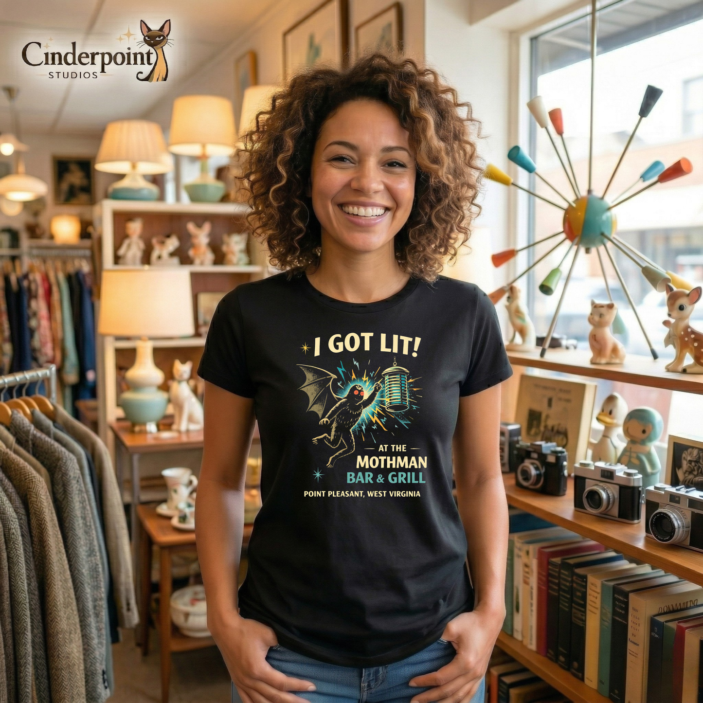 A woman in a vintage store is wearing a Black retro graphic t-shirt featuring Mothman flying into a glowing bug light with the phrase I Got Lit at the Mothman Bar and Grill, Point Pleasant, West Virginia.