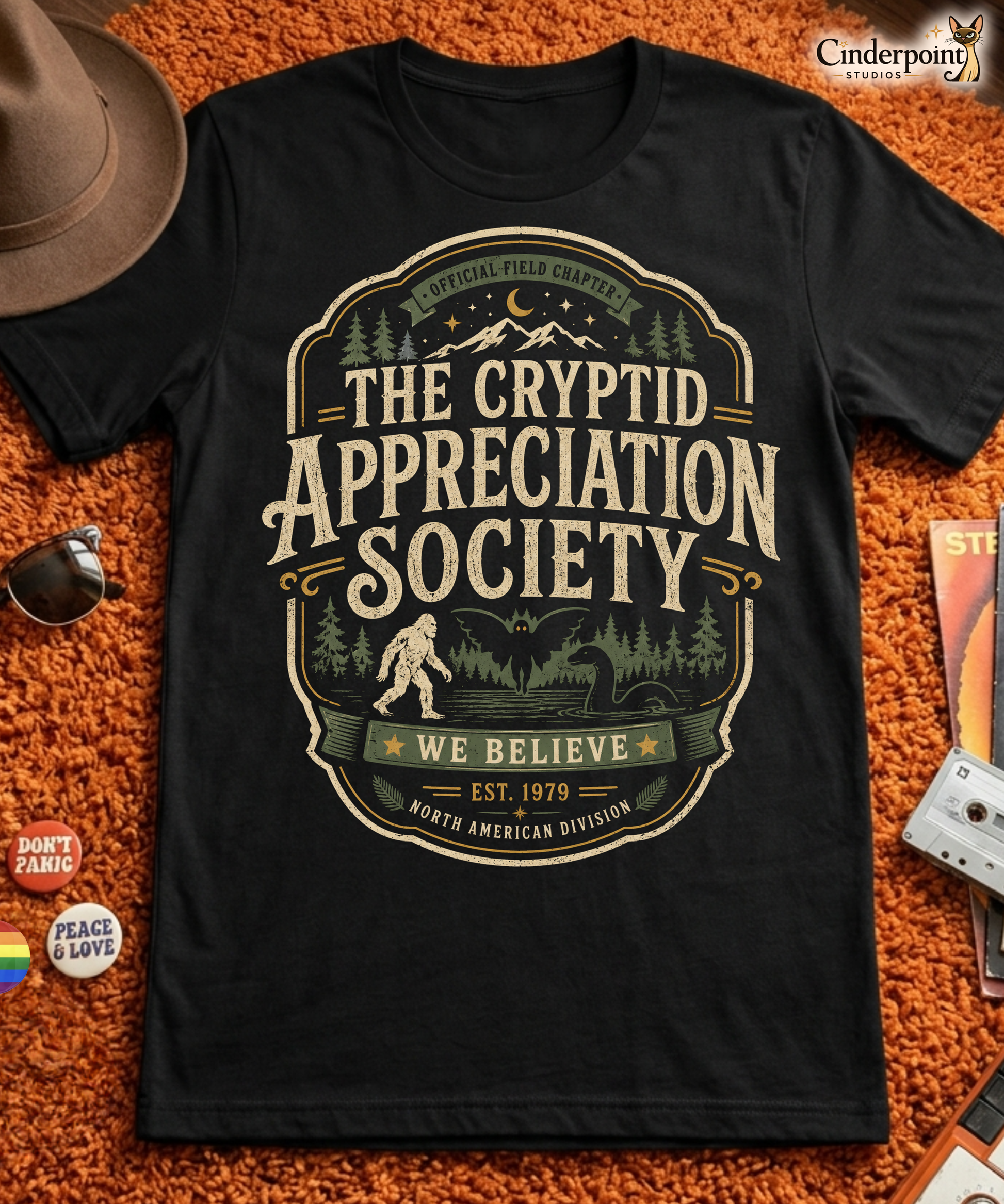 Black graphic tee featuring original Cinderpoint Studios artwork for The Cryptid Appreciation Society, a retro cryptid badge design with Sasquatch, Mothman, a Nessie-style lake creature, forest, mountains, moon, stars, and We Believe text.
