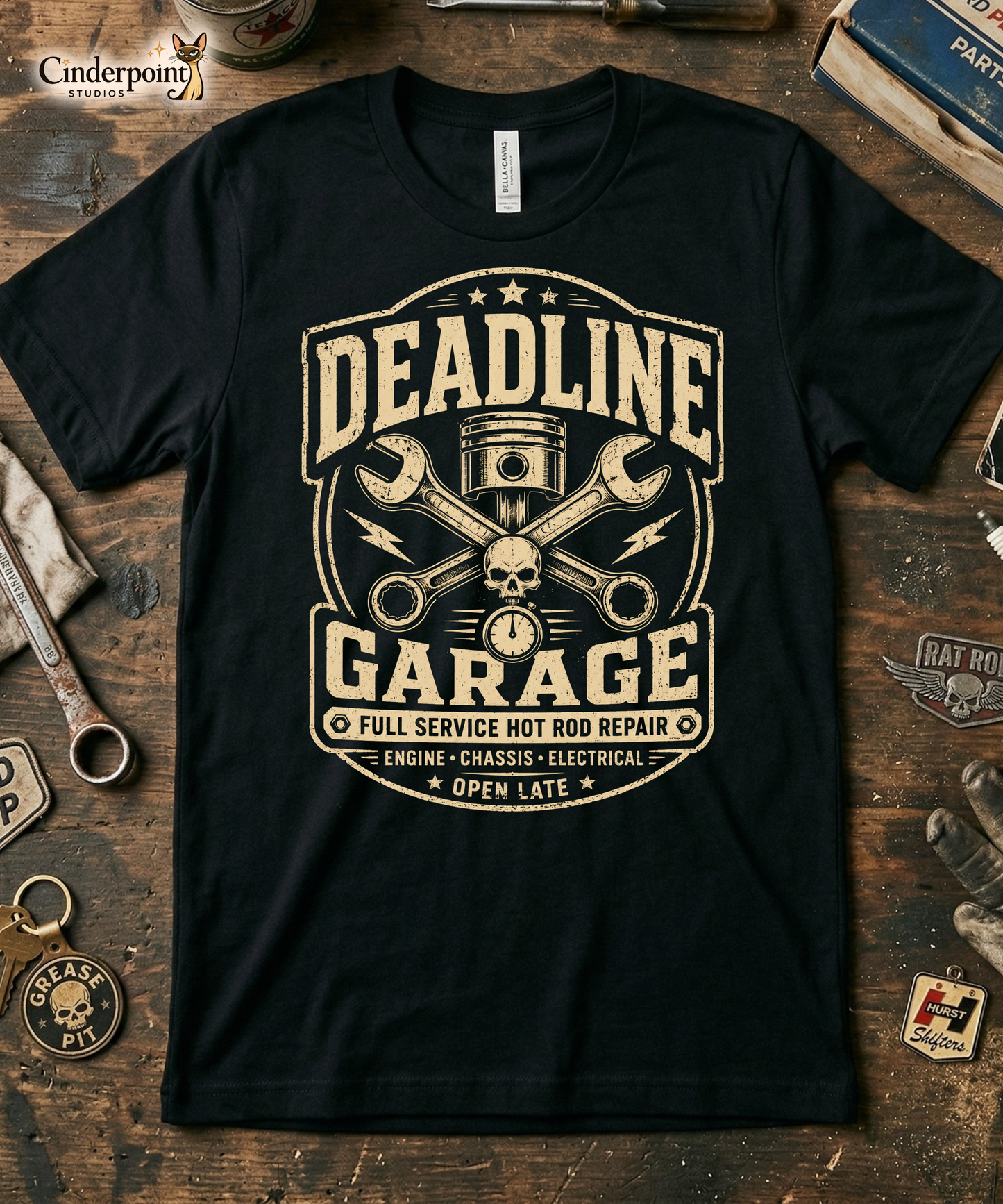 Mechanics workbench with a black graphic tee featuring original Cinderpoint Studios artwork for Deadline Garage, a one-color retro hot rod repair design with bold lettering, piston, crossed wrenches, skull detail, lightning bolts, stopwatch accent, and Full Service Hot Rod Repair text.
