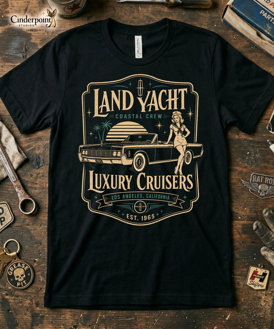 Workbench with a black graphic tee featuring original Cinderpoint Studios artwork for Land Yacht Luxury Cruisers, a retro classic car design with a 1960s luxury convertible inspired by the 1965 Continental, a sailor pinup, atomic accents, palm trees, and bold vintage lettering.