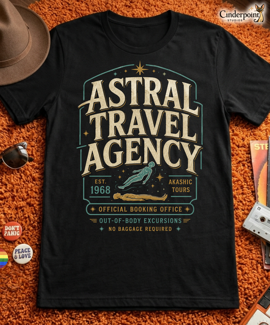 Black graphic tee featuring original Cinderpoint Studios artwork for Astral Travel Agency, a retro occult travel design with large vintage lettering, astral projection imagery, stars, and text including Official Booking Office, Akashic Tours, and Out-of-Body Excursions.