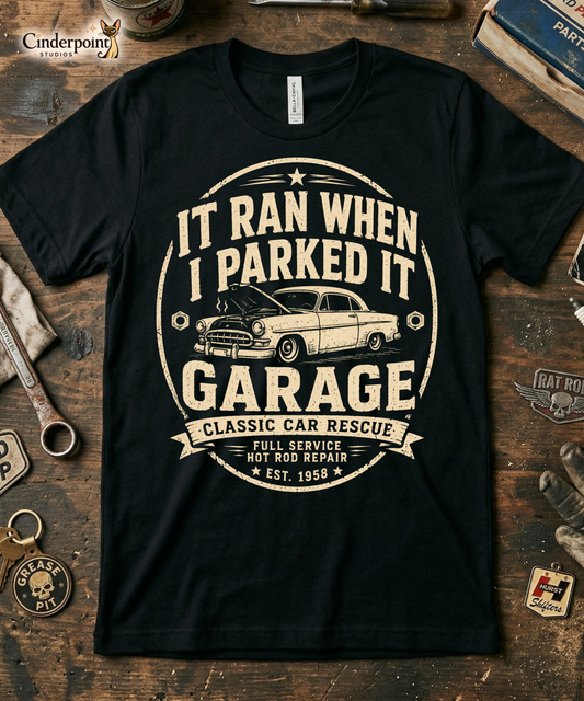 Workbench with a black graphic tee featuring original Cinderpoint Studios artwork for It Ran When I Parked It Garage, a one-color funny classic car design with bold lettering, a vintage car with the hood up, Classic Car Rescue text, and Full Service Hot Rod Repair details.