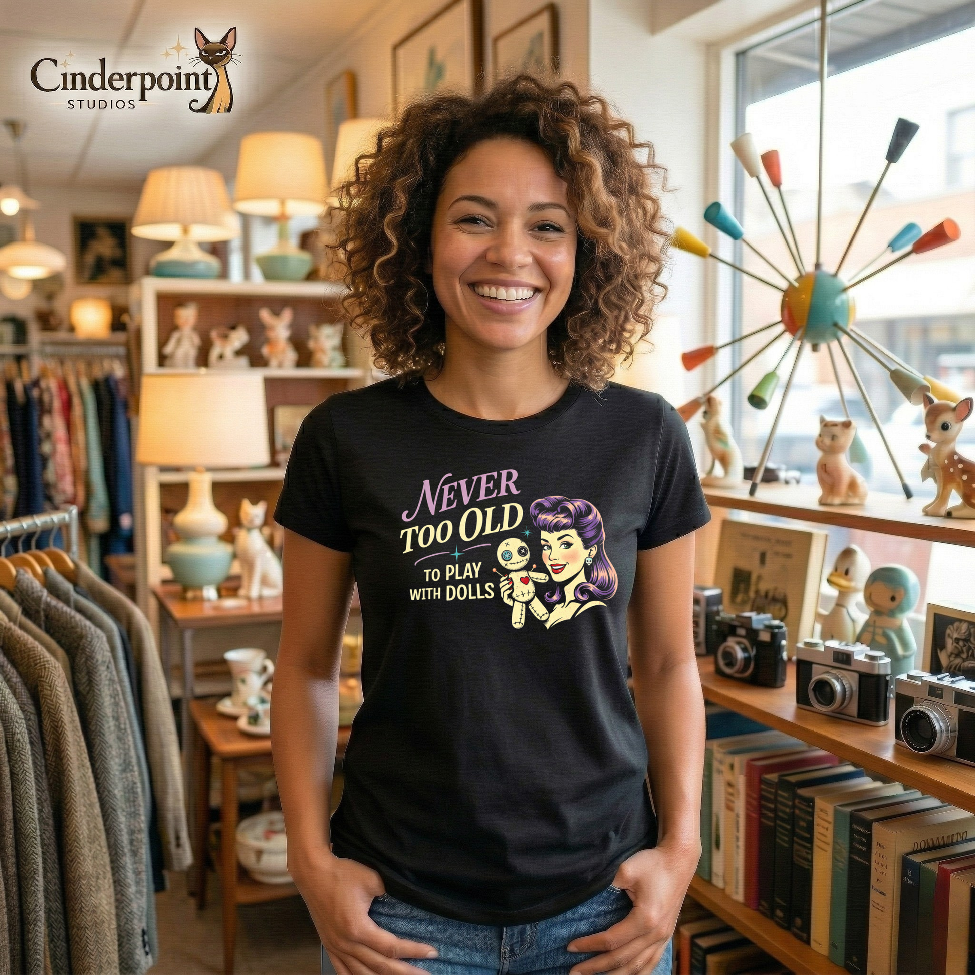 A woman in a vintage shop is wearing a Black retro graphic t-shirt featuring the phrase Never Too Old to Play with Dolls beside a vintage-inspired woman with purple hair holding a stitched voodoo doll.