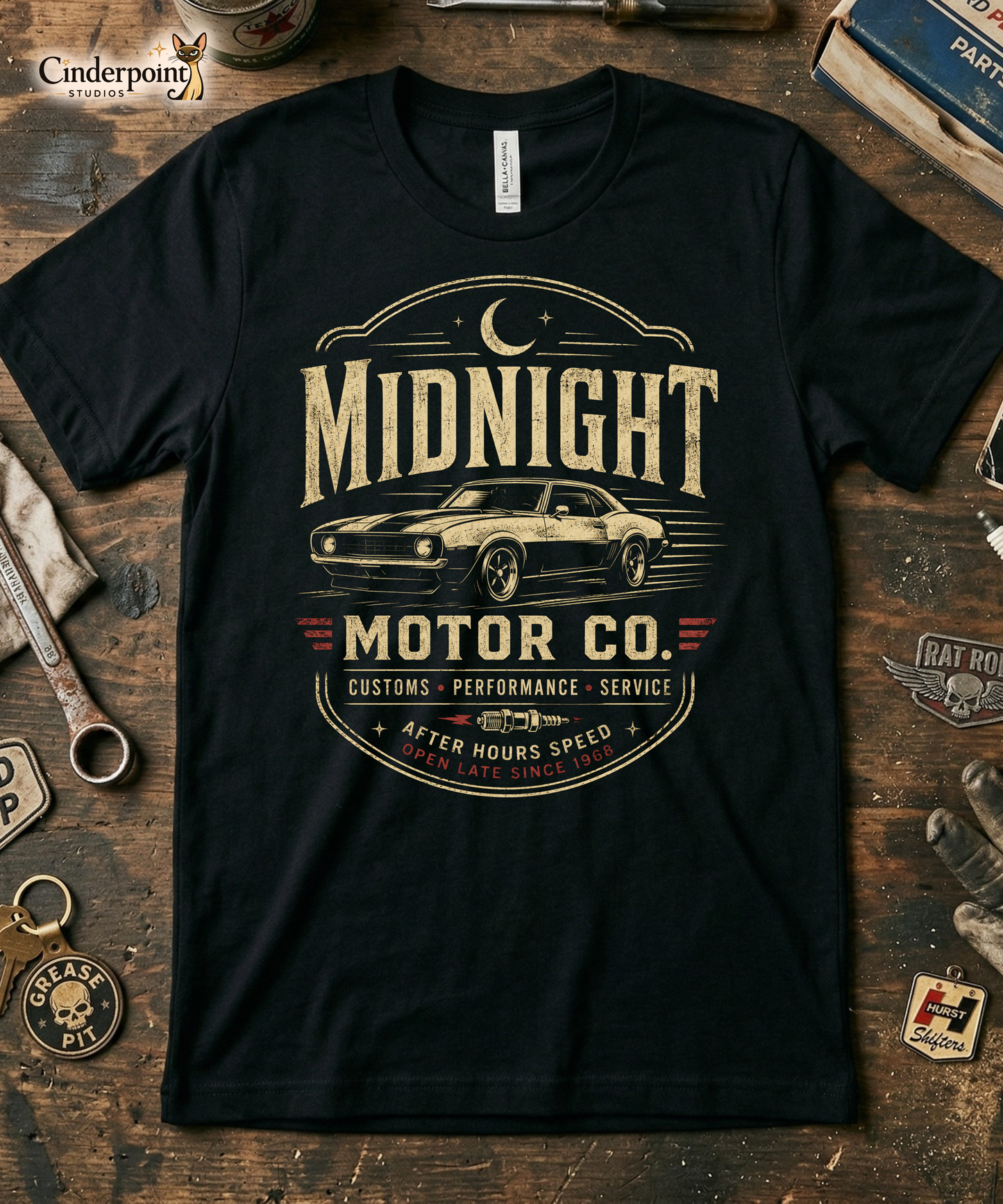 A workbench with a black graphic tee featuring original Cinderpoint Studios artwork for Midnight Motor Co., a retro hot rod garage design with a classic muscle car, crescent moon, spark plug detail, vintage lettering, red accent stripes, and After Hours Speed text.
