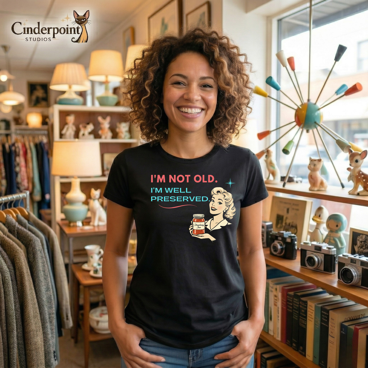 A lifestyle of a woman in a vintage shop, wearing a black retro graphic t-shirt featuring a smiling vintage-inspired woman holding a jar of strawberry jam with the phrase I’m Not Old, I’m Well Preserved in coral and aqua lettering.