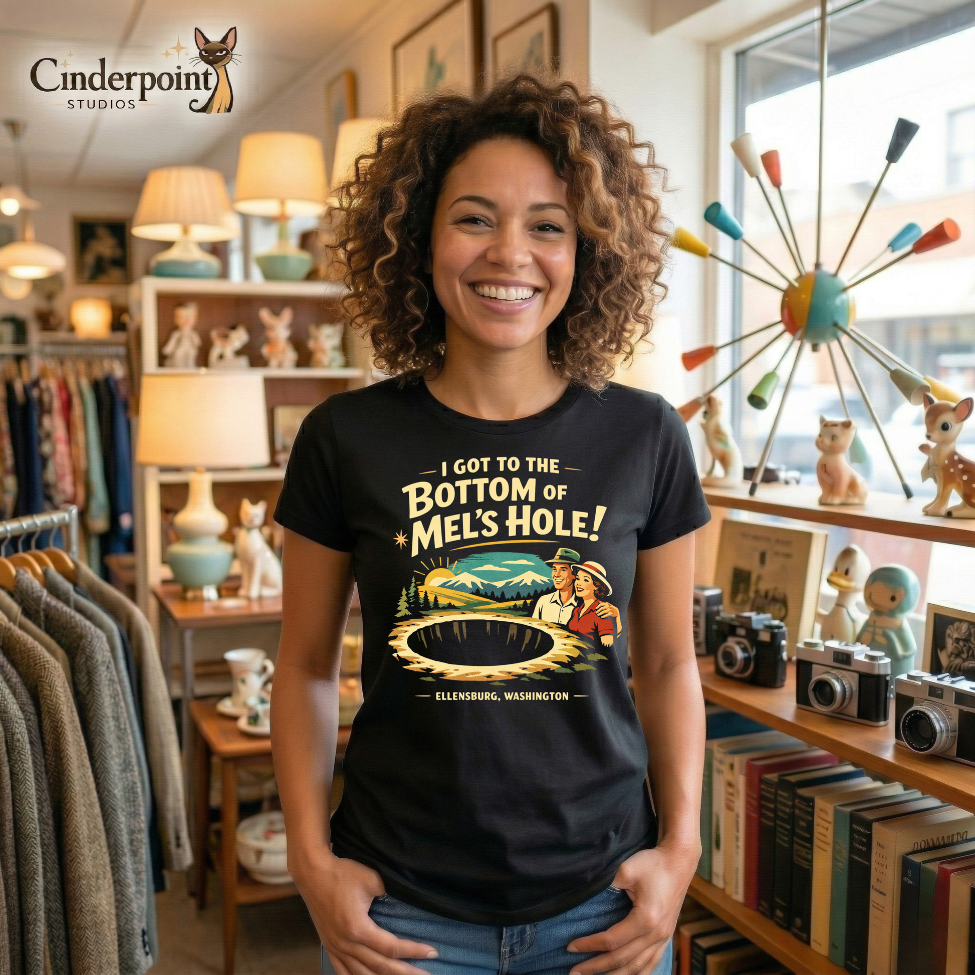 Woman in a vintage store wearing a Black retro graphic t-shirt featuring a vintage travel-style illustration of smiling tourists beside a giant mysterious hole with the phrase I Got to the Bottom of Mel’s Hole and Ellensburg, Washington.