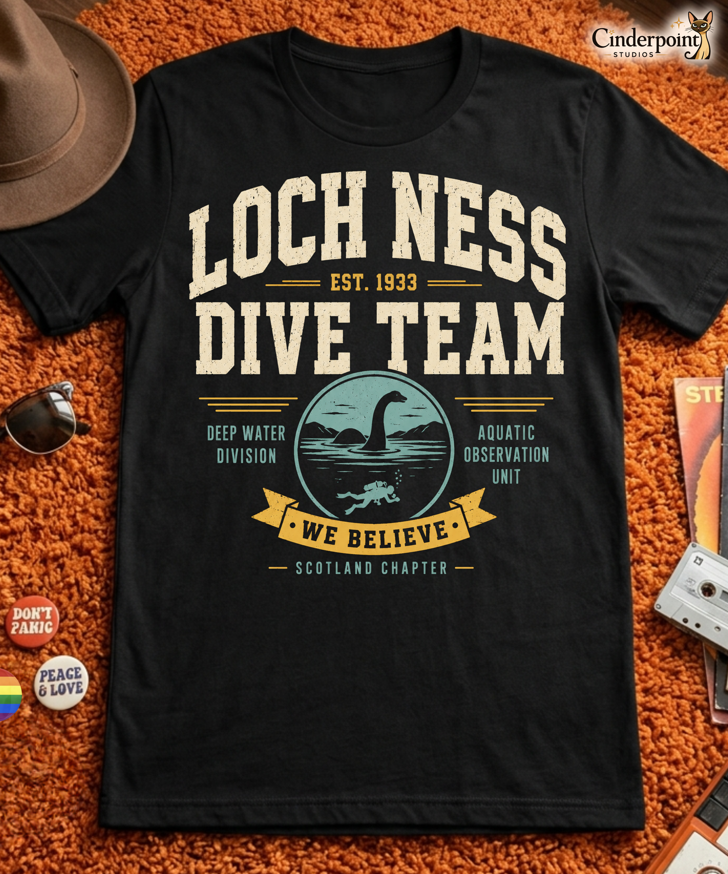 Black graphic tee featuring original Cinderpoint Studios artwork for Loch Ness Dive Team, a retro cryptid design with bold varsity lettering, a Nessie-style lake monster, diver, water scene, and text including Deep Water Division, Aquatic Observation Unit, We Believe, and Scotland Chapter.