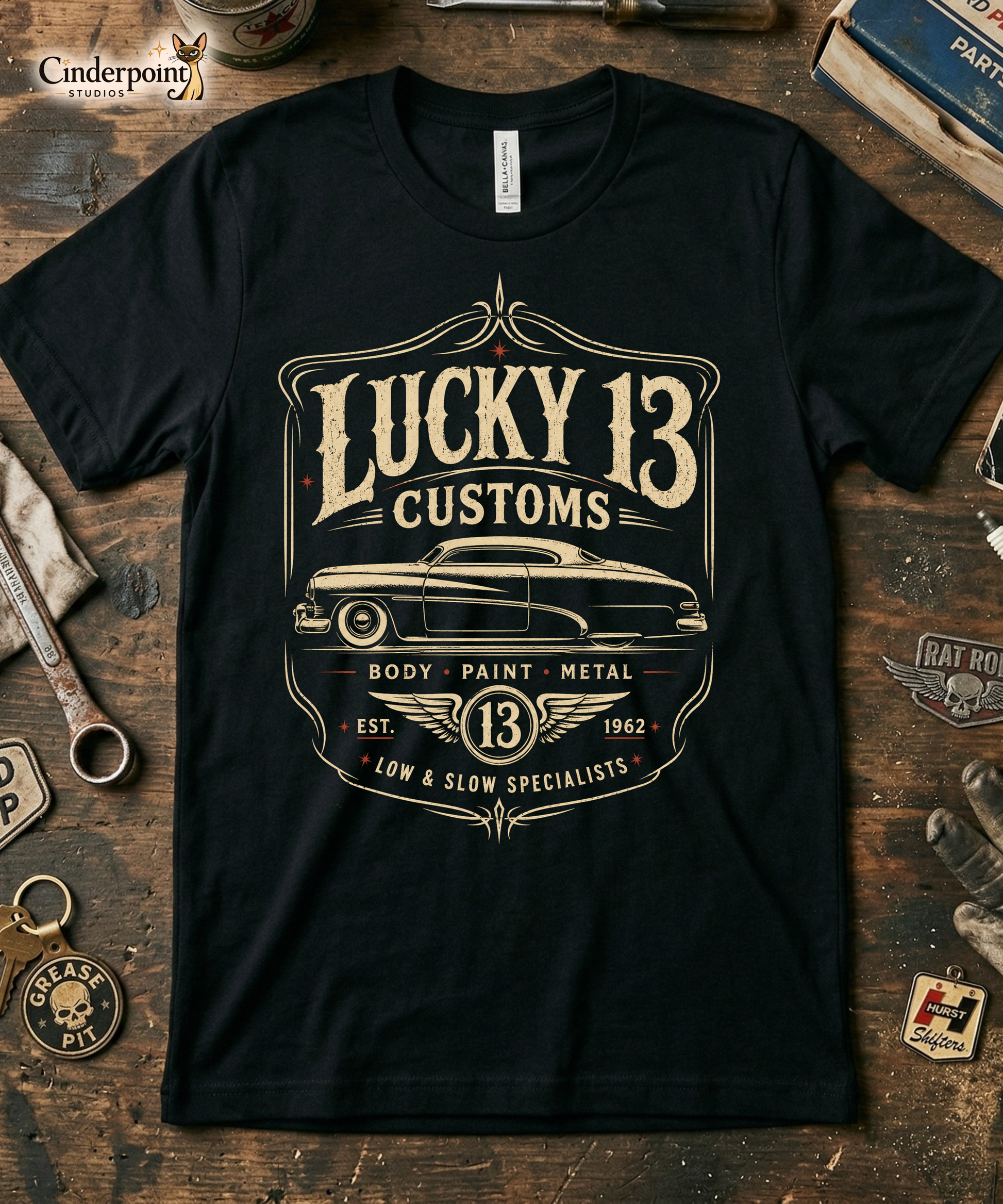Workbench with a black graphic tee featuring original Cinderpoint Studios artwork for Lucky 13 Customs, a retro custom shop design with a lowrider-style classic car, bold vintage lettering, winged 13 emblem, pinstripe-style frame, and Low & Slow Specialists text.