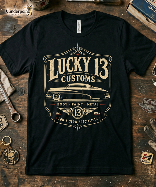 Workbench with a black graphic tee featuring original Cinderpoint Studios artwork for Lucky 13 Customs, a retro custom shop design with a lowrider-style classic car, bold vintage lettering, winged 13 emblem, pinstripe-style frame, and Low & Slow Specialists text.