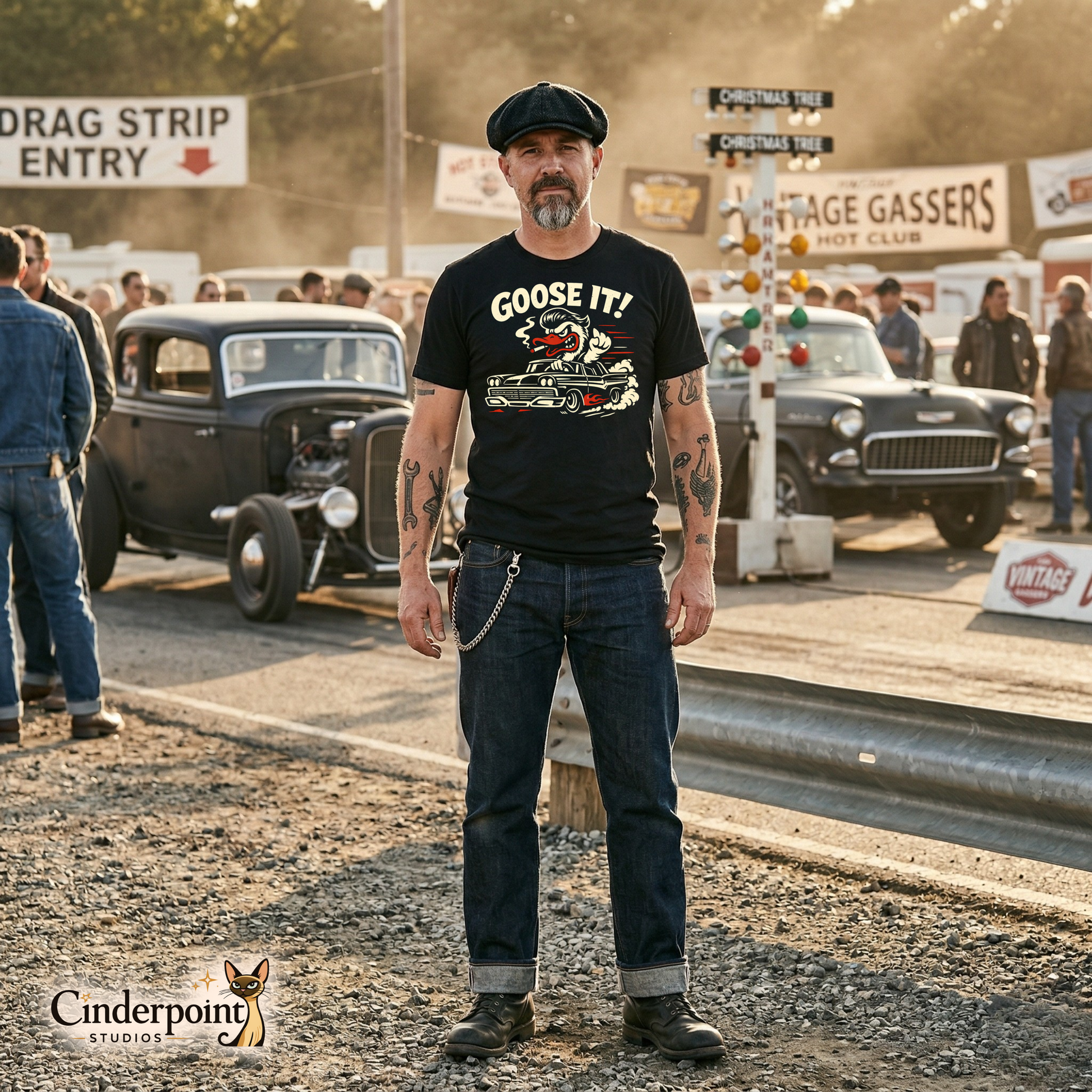 A man in a flat cap and tattoos wearing the black "Goose It!" t-shirt standing at a vintage drag strip entry with hot rods blurred in the background.