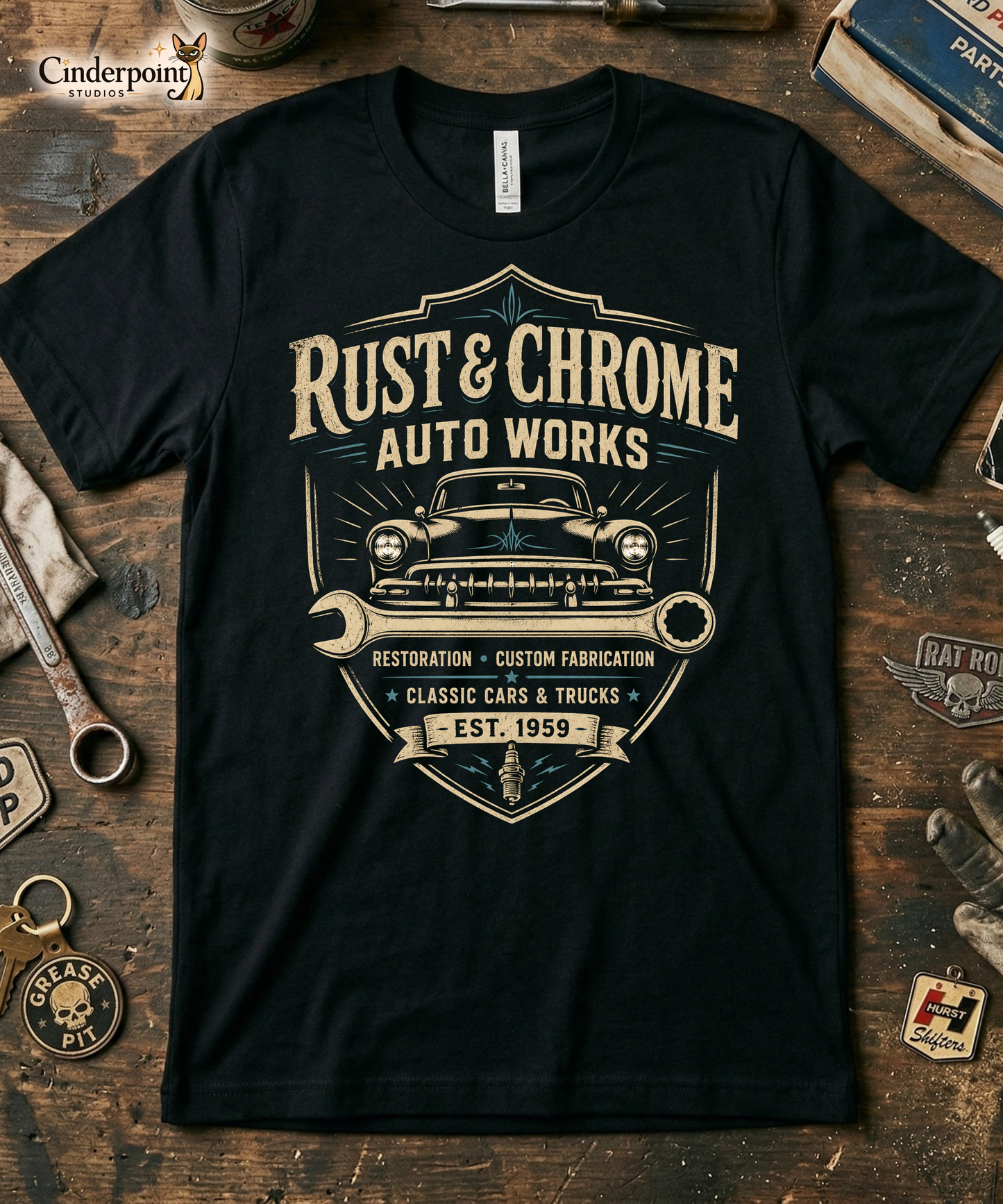 Workbench with a black graphic tee featuring original Cinderpoint Studios artwork for Rust & Chrome Auto Works, a vintage garage design with a front-facing classic car, bold lettering, wrench accent, spark plug detail, restoration and custom fabrication text, and shield-style framing.
