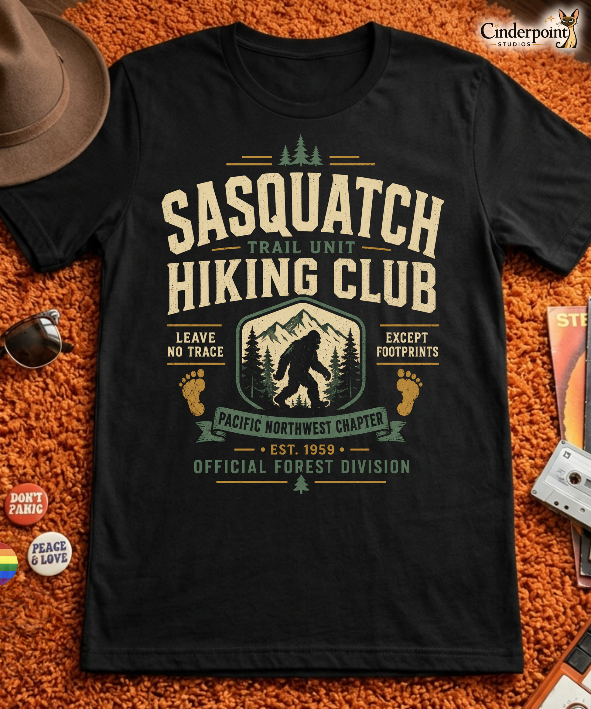 Black graphic tee featuring original Cinderpoint Studios artwork for Sasquatch Hiking Club, a retro Bigfoot trail design with bold varsity lettering, mountain scenery, pine trees, footprint accents, and text including Trail Unit, Pacific Northwest Chapter, Leave No Trace, Except Footprints, and Official Forest Division.