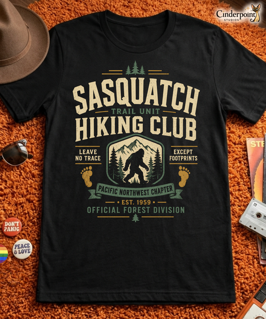 Black graphic tee featuring original Cinderpoint Studios artwork for Sasquatch Hiking Club, a retro Bigfoot trail design with bold varsity lettering, mountain scenery, pine trees, footprint accents, and text including Trail Unit, Pacific Northwest Chapter, Leave No Trace, Except Footprints, and Official Forest Division.
