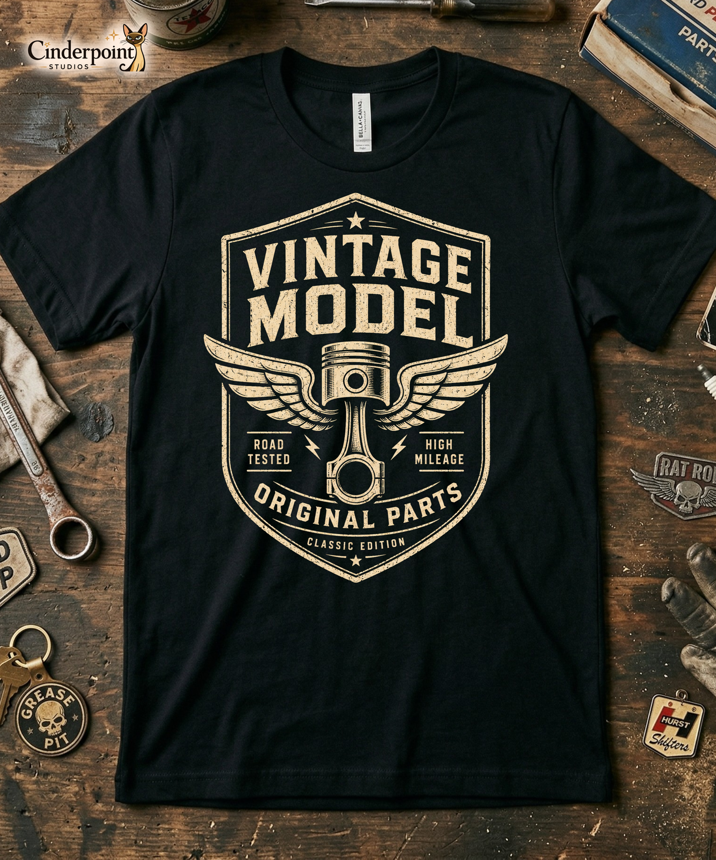 Black graphic tee featuring original Cinderpoint Studios artwork for Vintage Model, a one-color retro garage badge design with bold lettering, a winged piston, and text including Road Tested, High Mileage, Original Parts, and Classic Edition.
