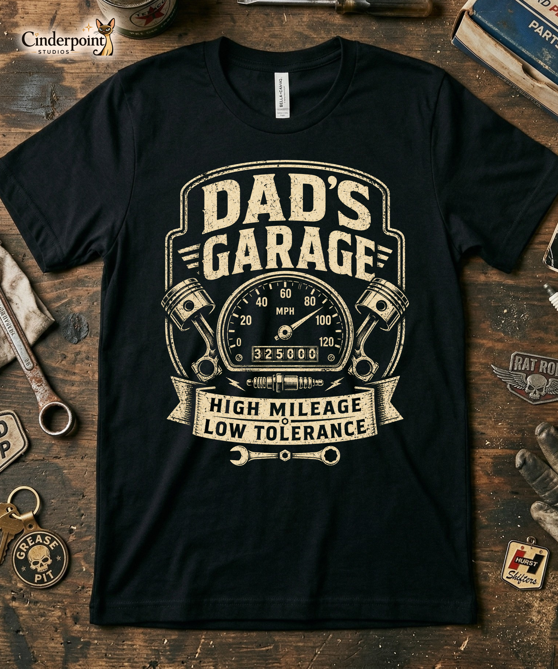 Black graphic tee featuring original Cinderpoint Studios artwork for Dad’s Garage, a one-color retro garage badge design with bold lettering, a speedometer, pistons, spark plug, wrench, and High Mileage Low Tolerance text.